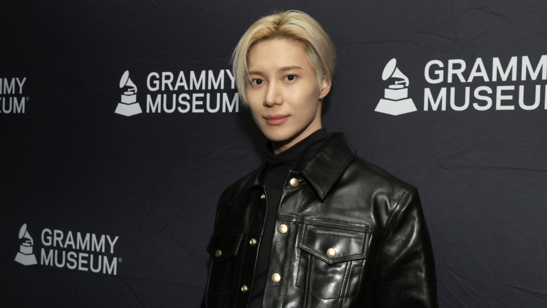 K-pop superstar Taemin with blonde hair in a black leather jacket stands in front of a "GRAMMY MUSEUM" backdrop.