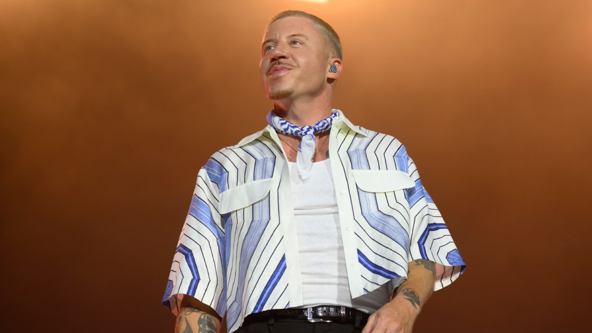 Macklemore on stage wearing a striped shirt and bandana, with tattoos visible, stands confidently under warm lighting.