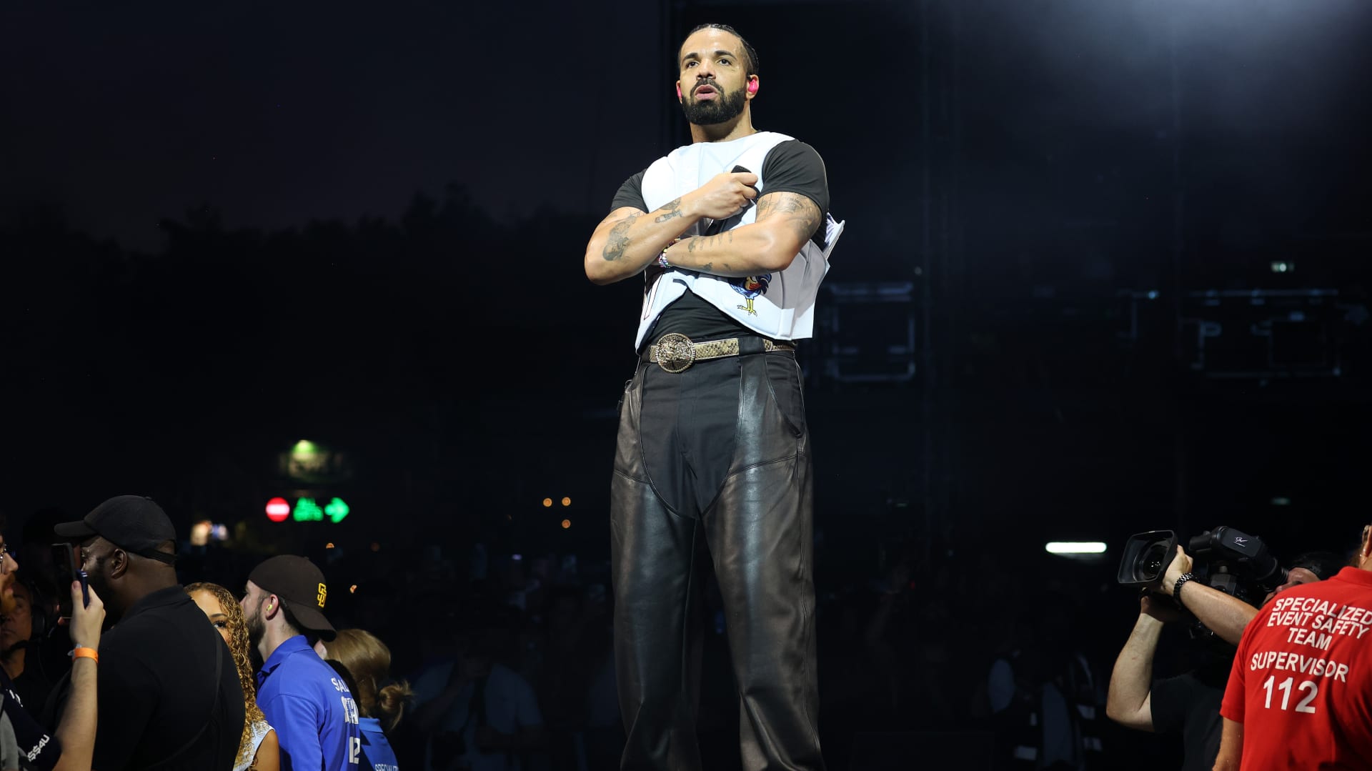 Drake stands on stage wearing a white and black shirt and leather pants, with people and security staff in the background.
