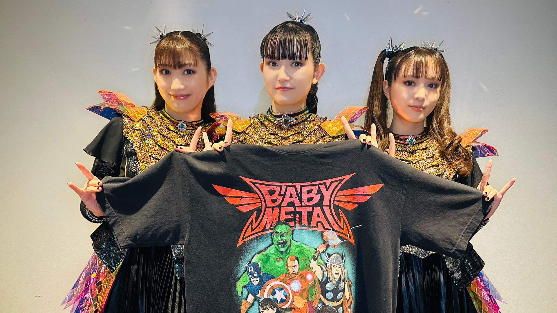 Three members of Babymetal in colorful costumes hold a Babymetal T-shirt with superhero graphics, smiling and making hand gestures.