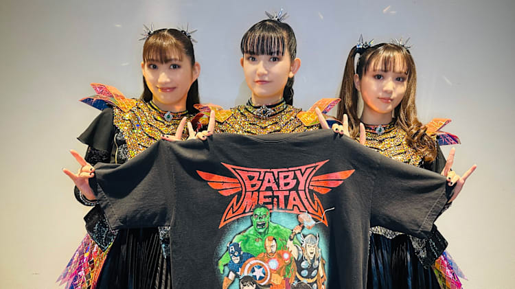 Three members of Babymetal in colorful costumes hold a Babymetal T-shirt with superhero graphics, smiling and making hand gestures.