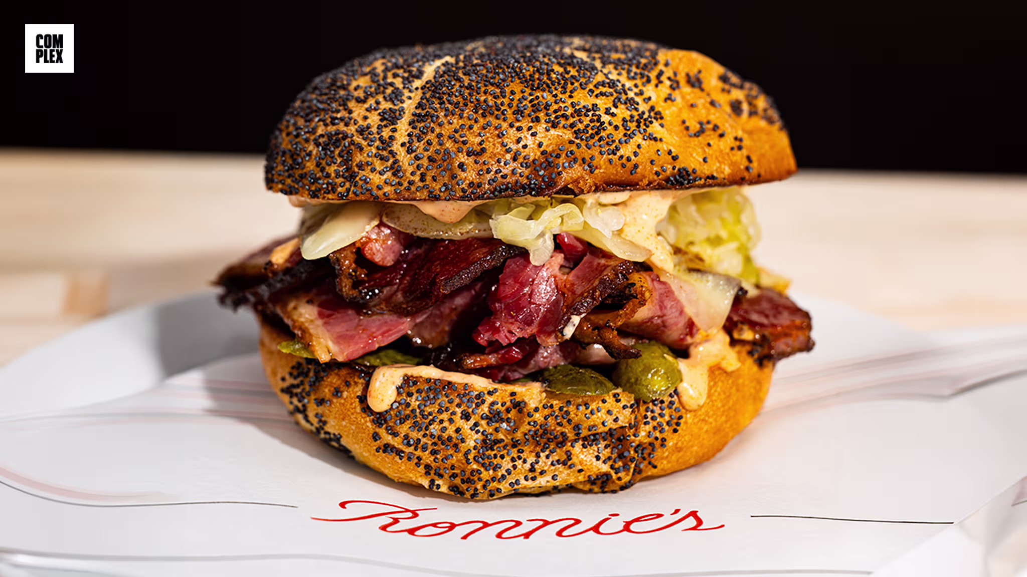 A poppy seed bun sandwich filled with sliced meat, pickles, and cabbage on a plate with "Ronnie's" written on it.