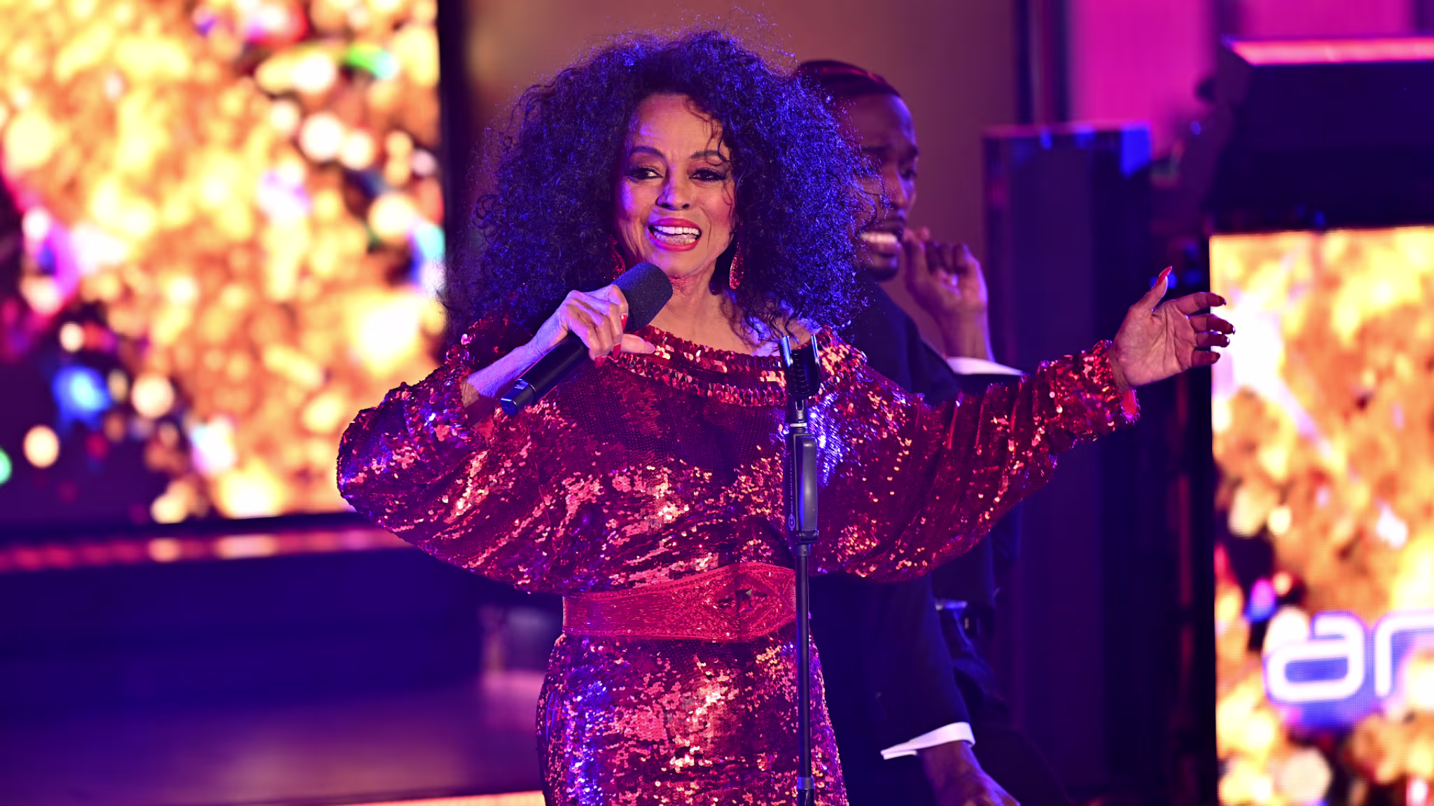 Diana Ross performing on stage in a sparkling red dress, holding a microphone, with a vibrant background.