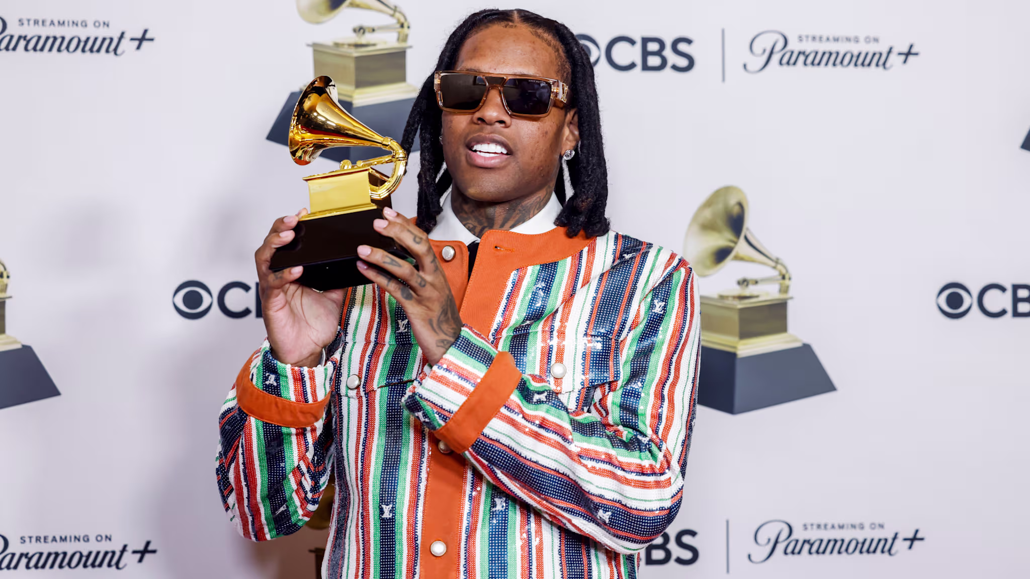 Lil Durk in a colorful striped jacket holds a Grammy award, standing in front of a CBS and Paramount+ backdrop.