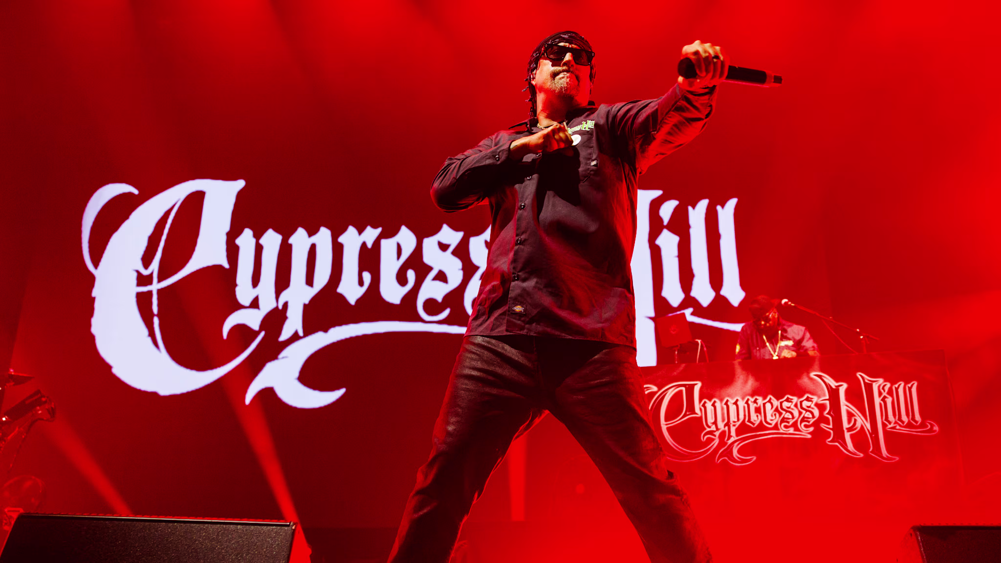Cypress Hill performing on stage with vibrant red lighting, their logo displayed prominently in the background.