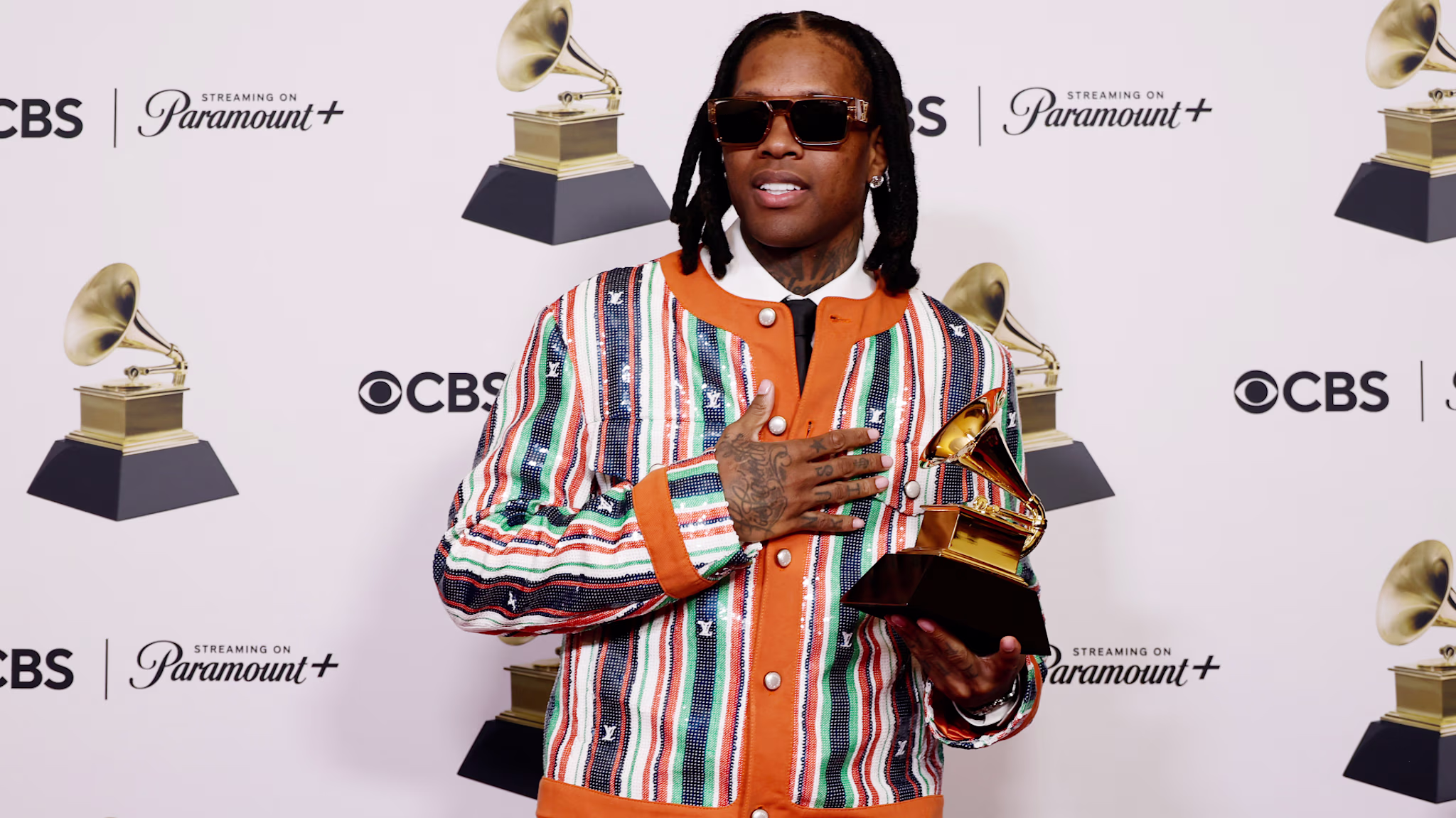 Lil Durk wearing sunglasses and a colorful jacket holds a Grammy award, standing in front of a backdrop with CBS and Paramount+ logos.