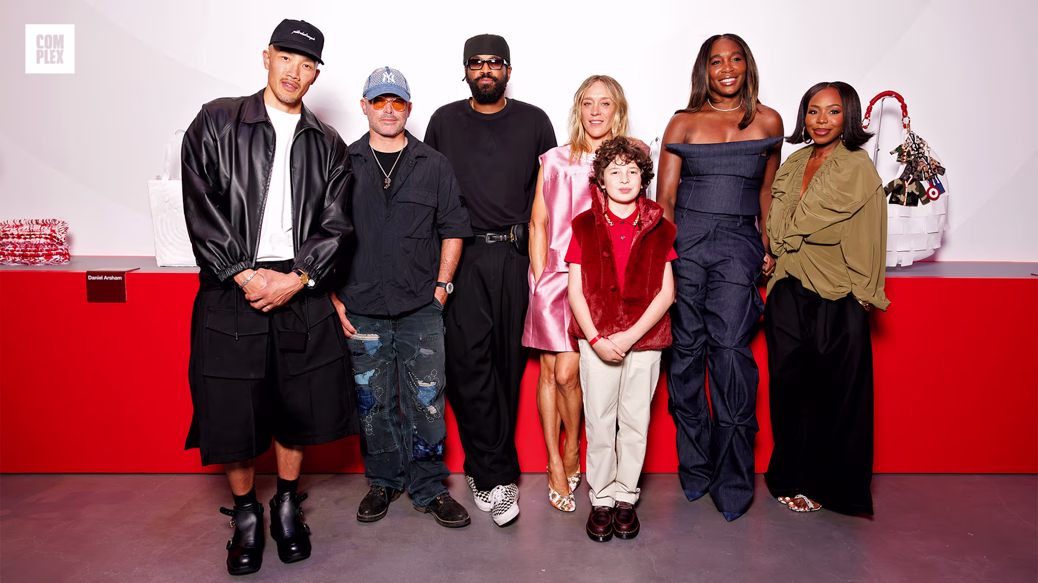 A group of people, including a child in a red outfit, stand in front of a red and white backdrop. They are dressed in stylish, modern clothing.