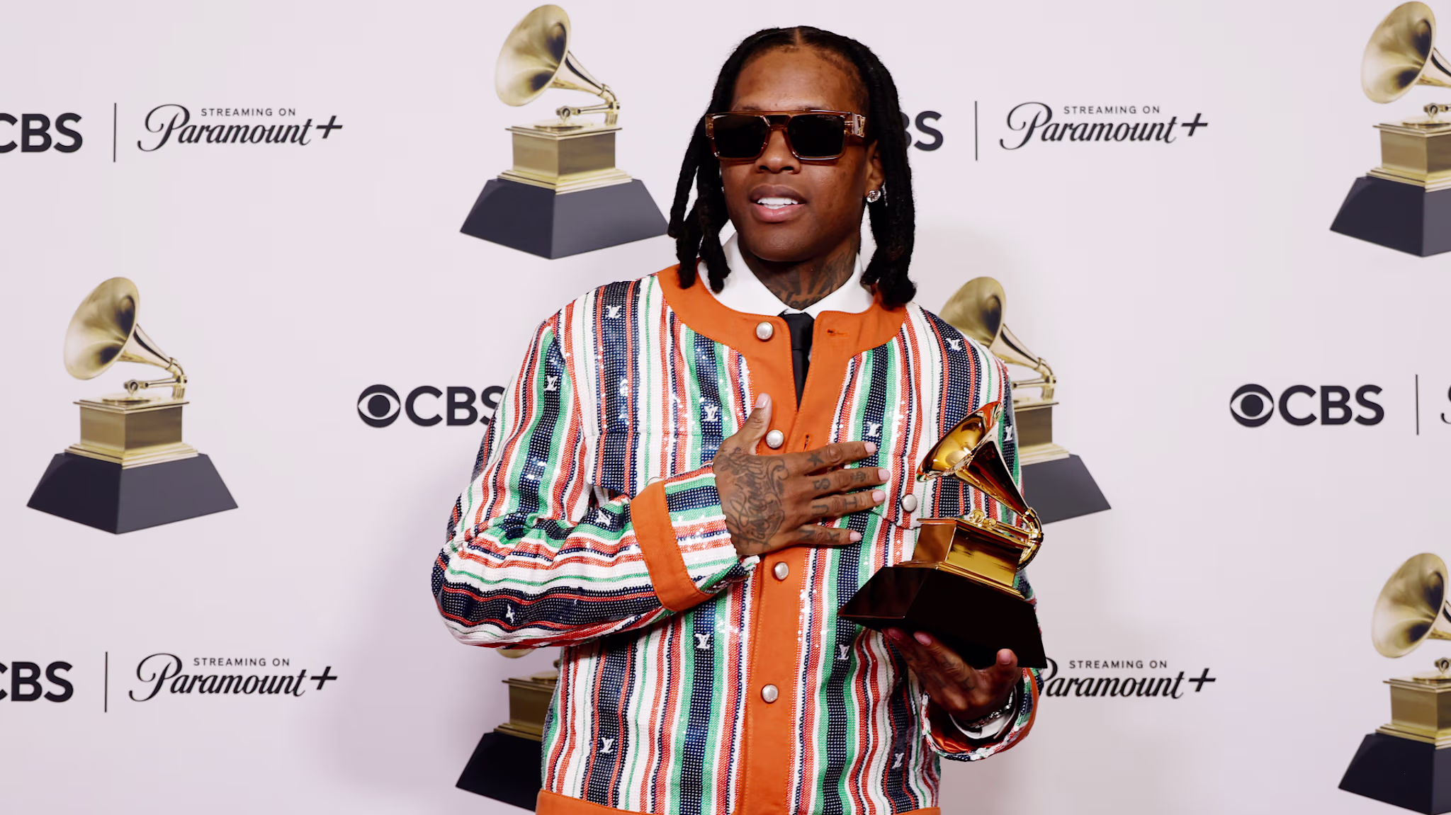 Durk wearing sunglasses and a colorful striped jacket holds a Grammy award, standing in front of a backdrop with CBS and Paramount+ logos.