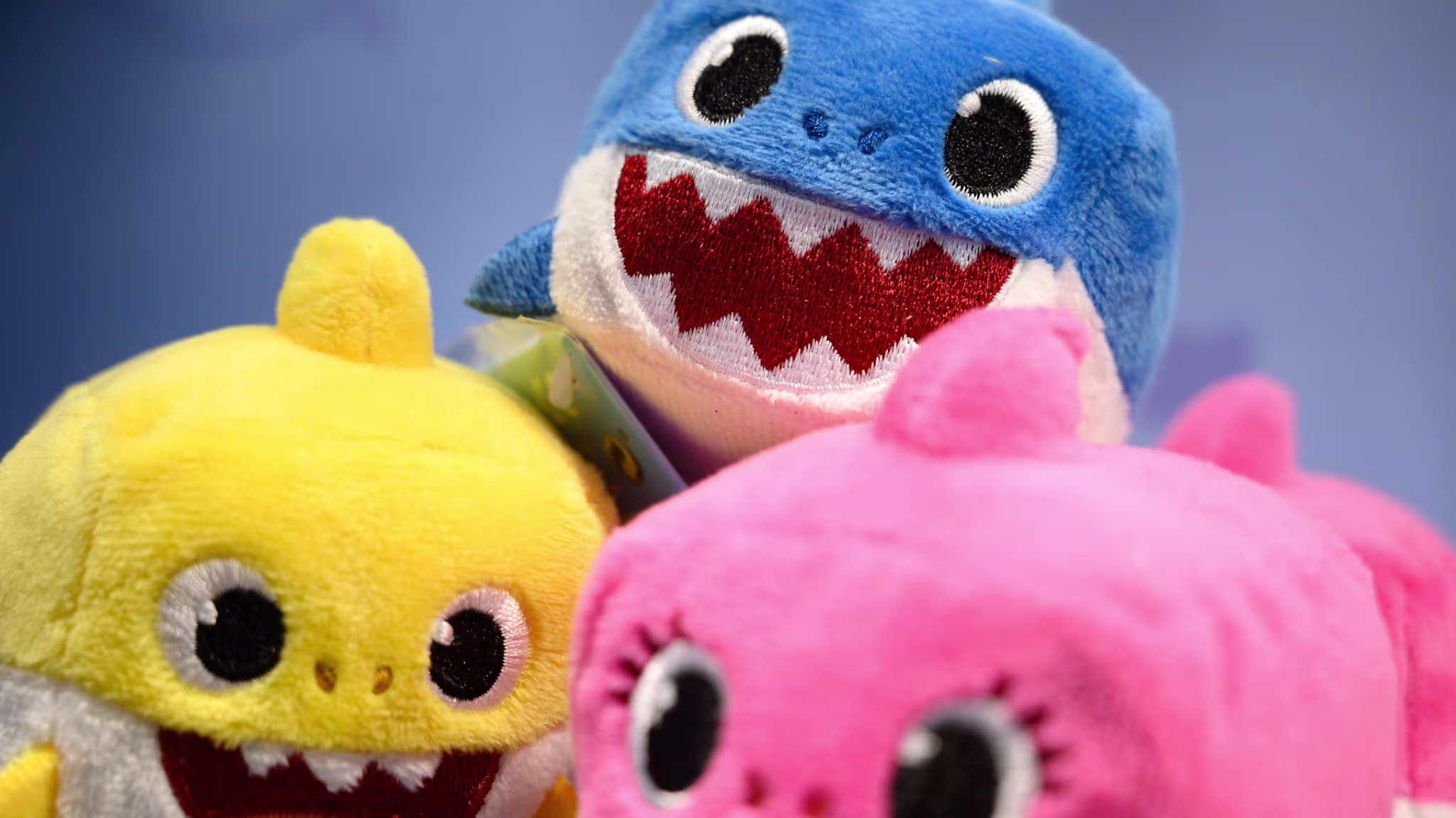 Three plush toys resembling sharks: one blue, one yellow, and one pink, with wide eyes and open mouths.