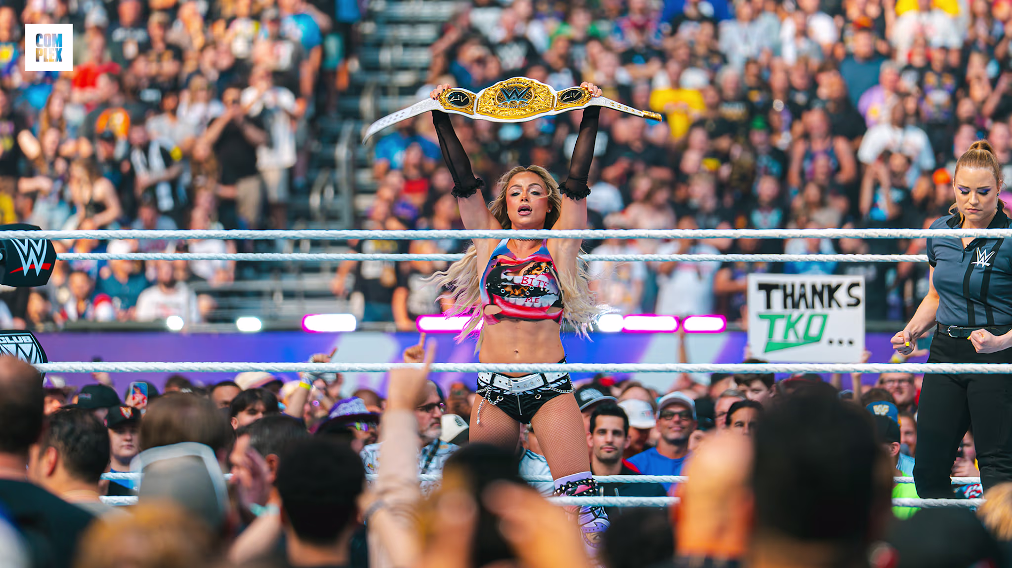 A female wrestler holds a championship belt in a ring, surrounded by a cheering crowd.