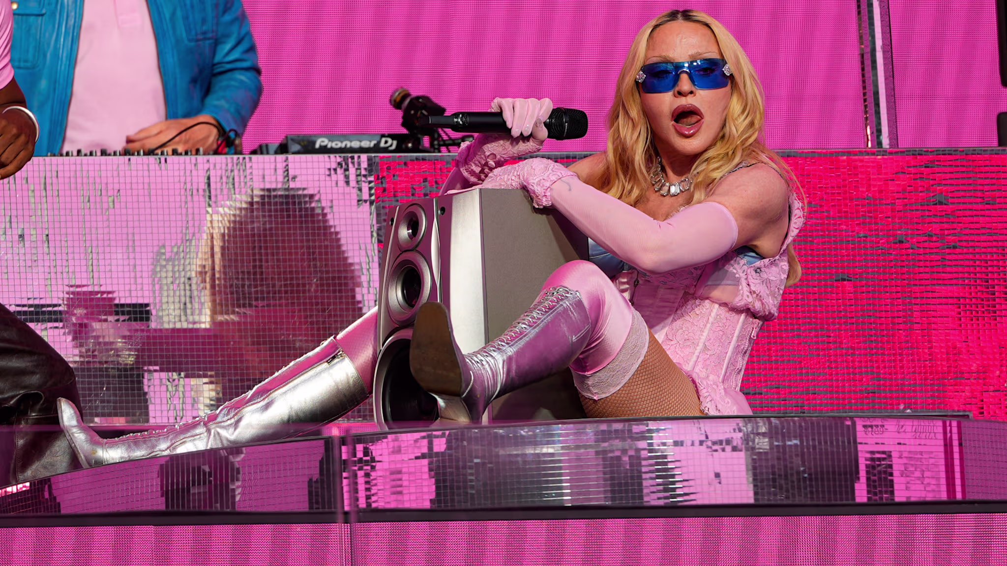 Madonna in a pink corset, silver boots, and blue sunglasses sings into a microphone on a pink stage.
