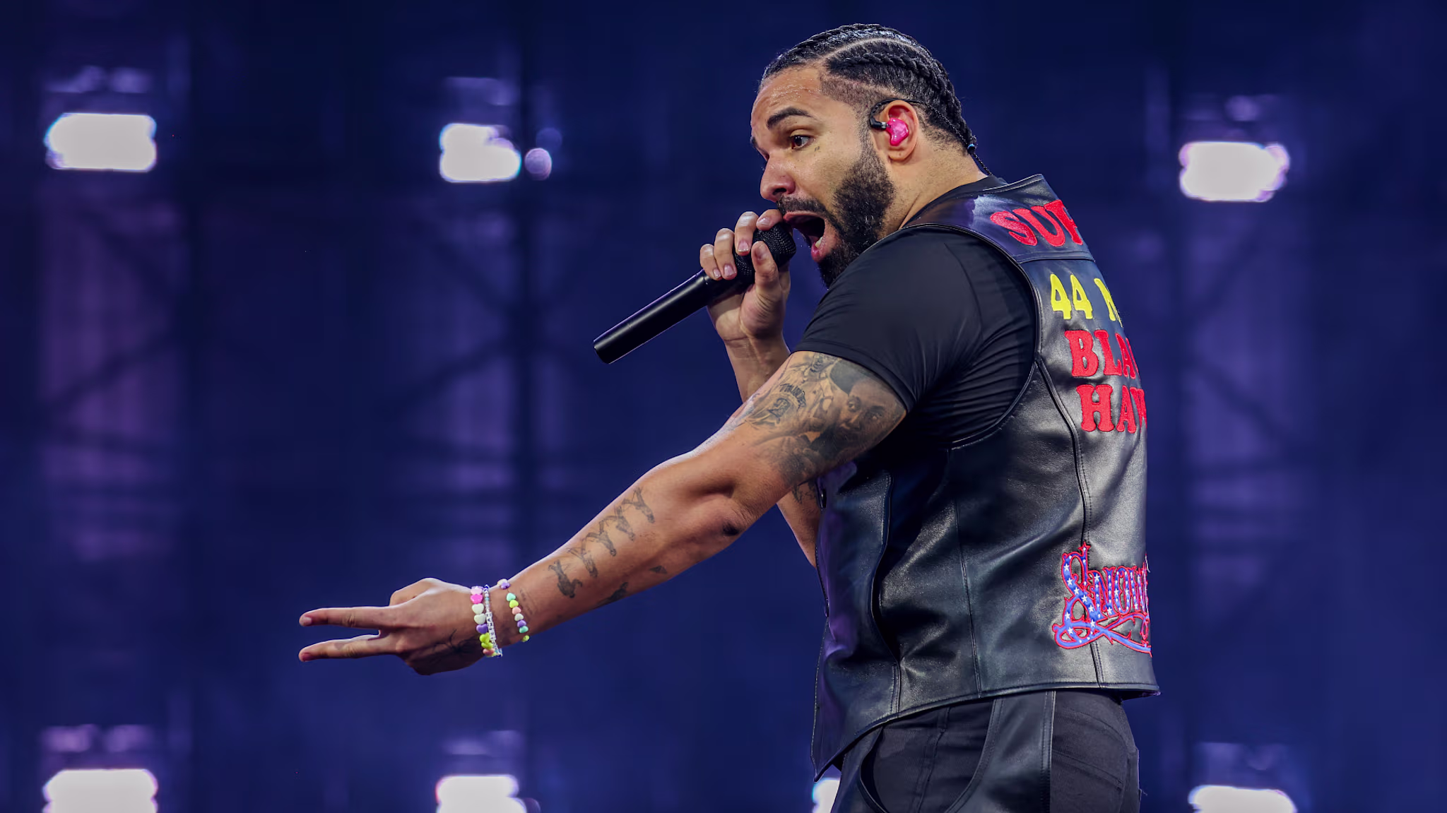 Drake performing on stage, wearing a black vest with colorful text, holding a microphone, and gesturing with his hand.