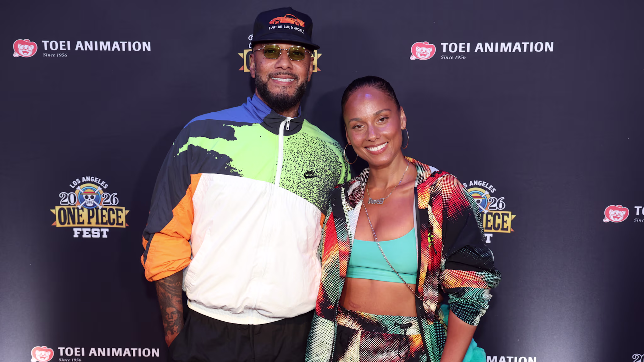 Alicia Keys and Swizz Beatz at an event, standing in front of a "One Piece Fest" backdrop, smiling and posing together.