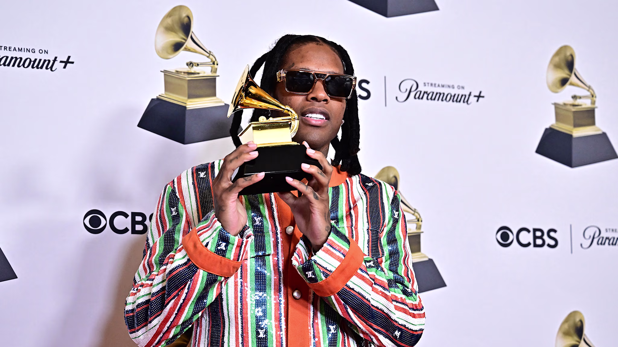 Lil Durk in a colorful striped jacket holds a Grammy award, posing on the red carpet with logos for CBS and Paramount+ in the background.
