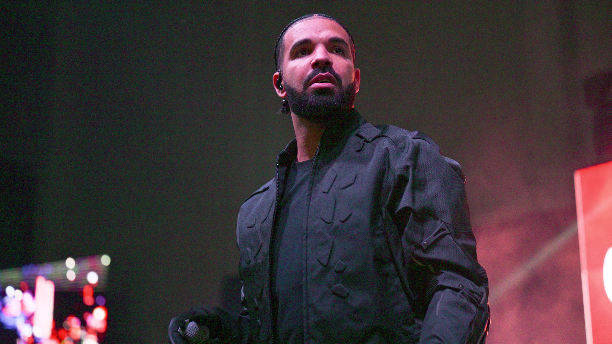 Drake performing on stage, wearing a black jacket, holding a microphone, with a focused expression.