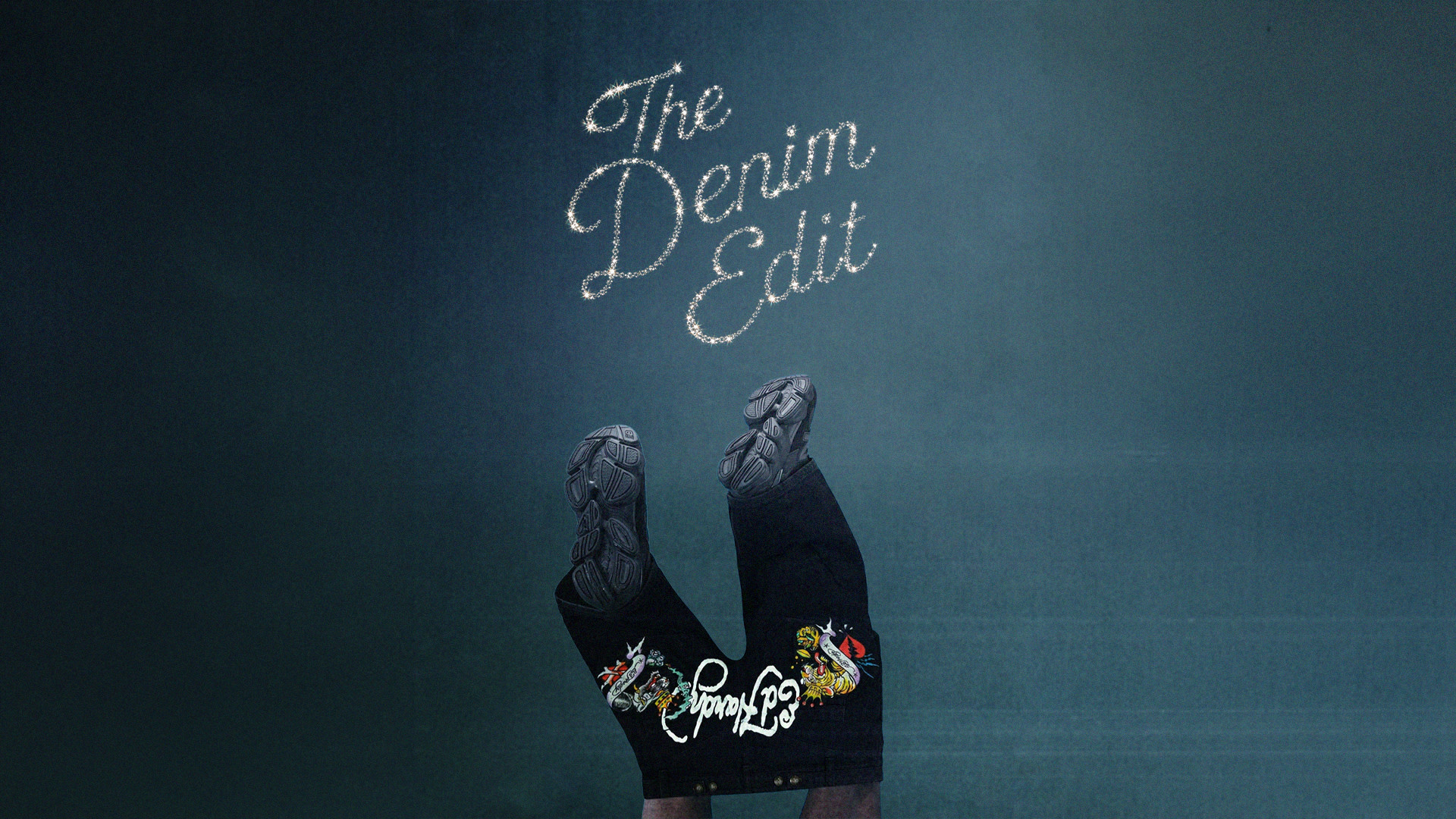 Legs wearing black jeans with colorful embroidery and sneakers, pointing upwards against a dark background with "The Denim Edit" text.