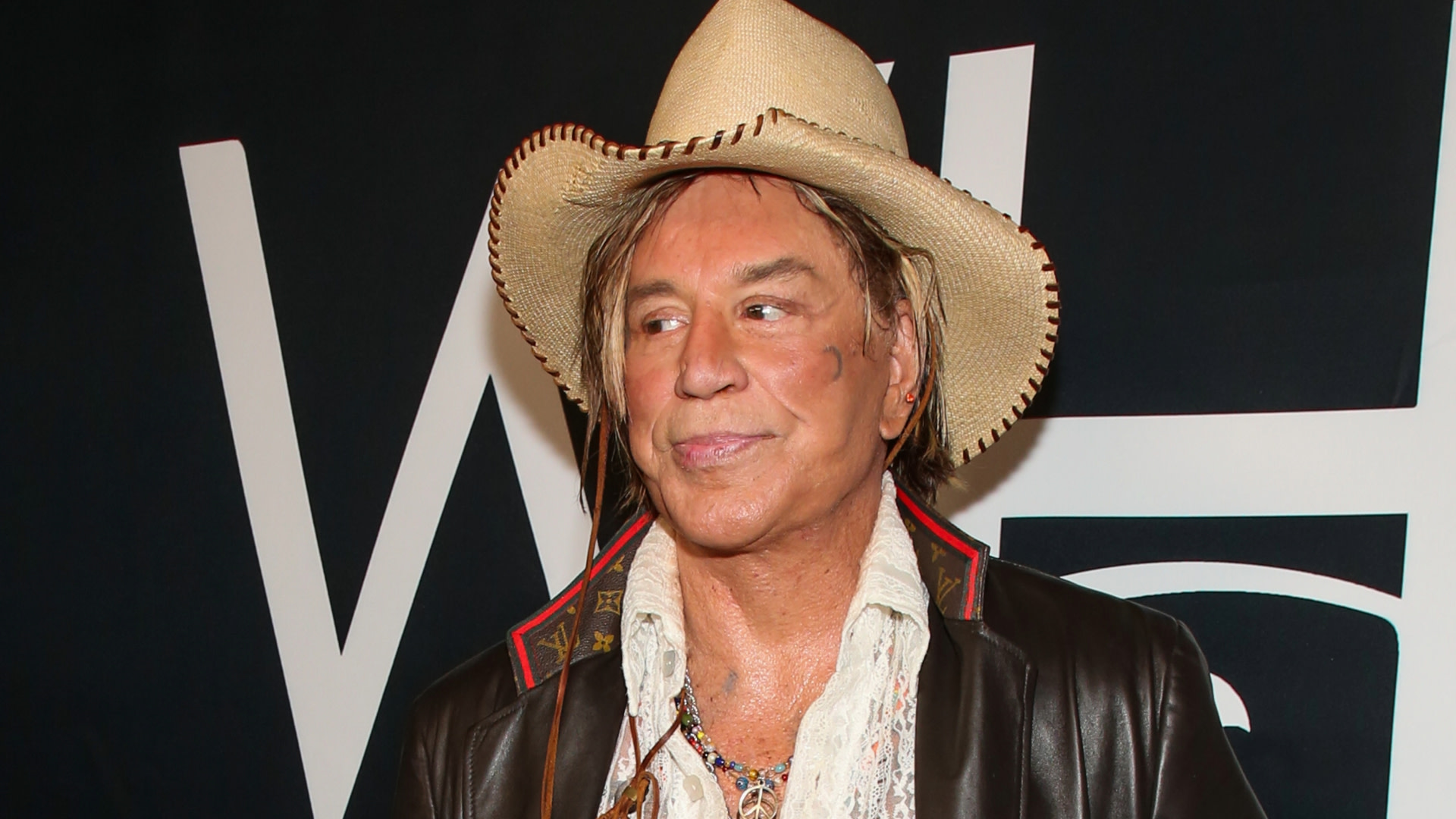 A man wearing a cowboy hat, leather jacket, and white shirt stands in front of a dark background with white letters.
