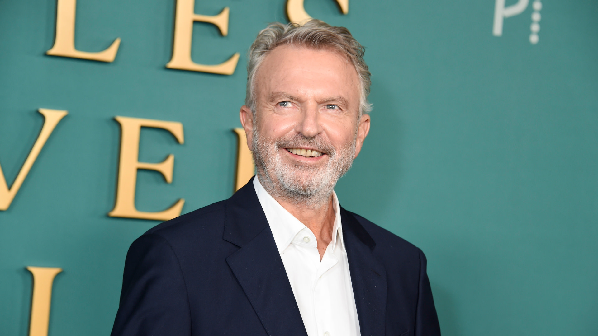 Sam Neill, wearing a dark suit and white shirt, smiles on a red carpet with a green background.