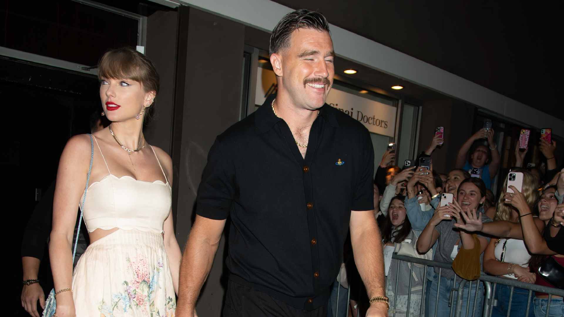 A woman in a white floral dress and a man in a black shirt walk past a crowd of fans taking photos.