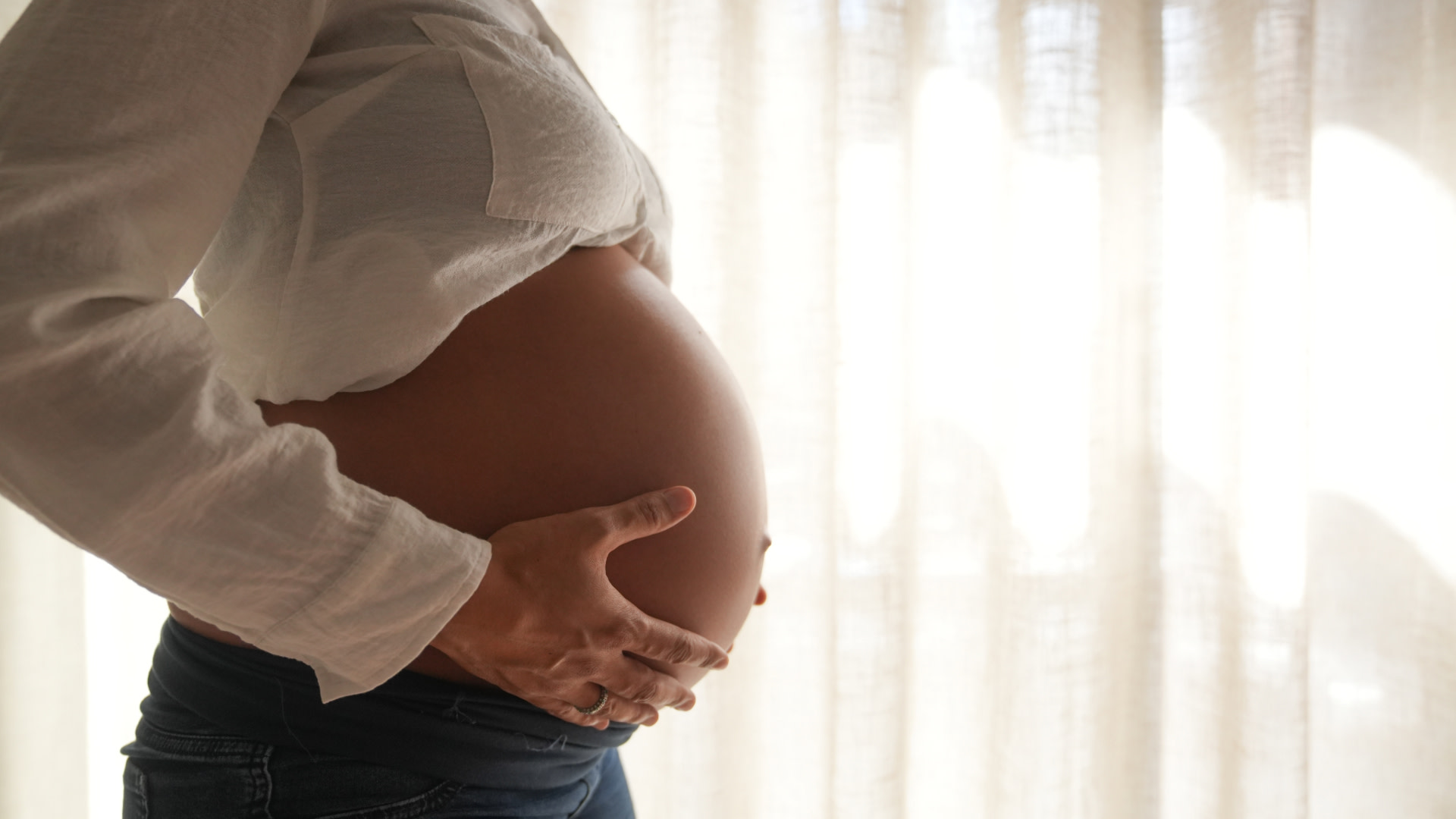 A pregnant woman in a white blouse holds her belly in profile, standing by a softly lit curtain.