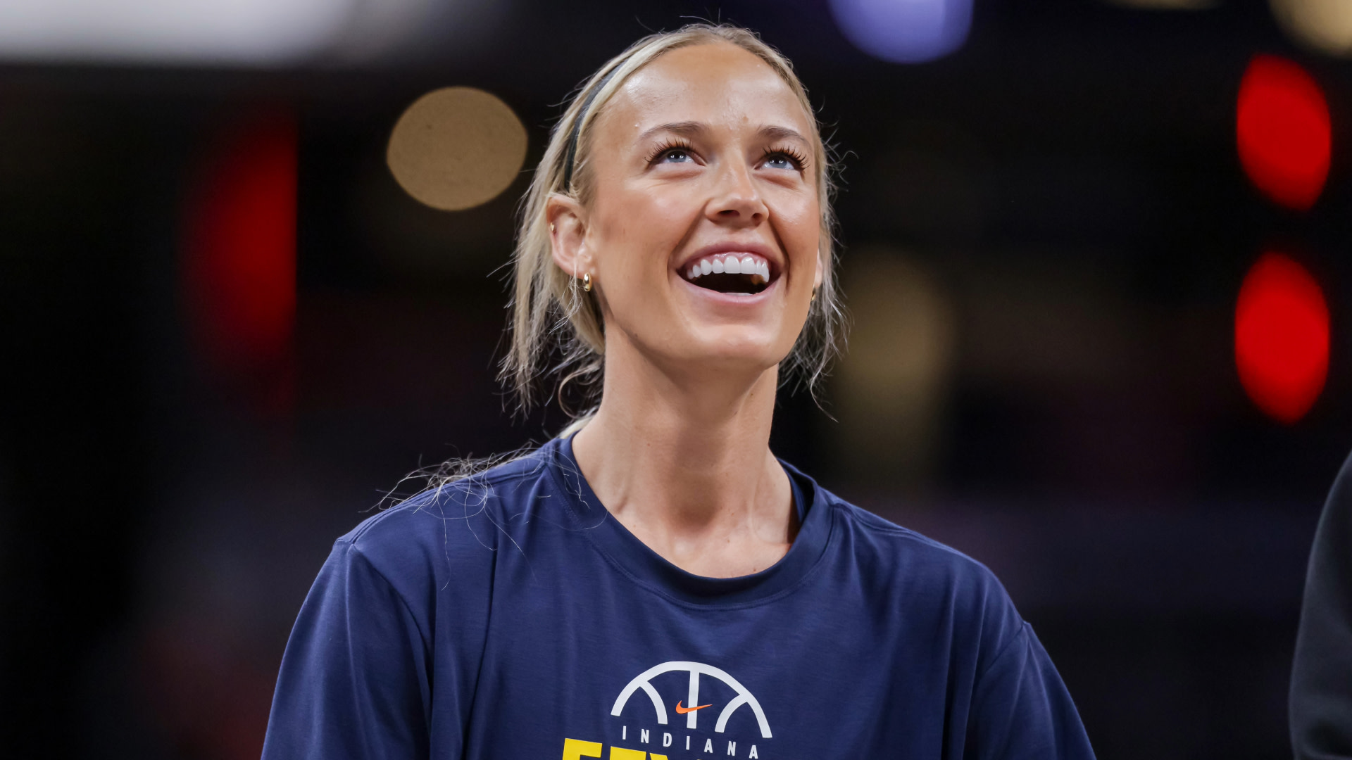 A smiling woman wearing an Indiana Fever shirt, looking upwards. The background is blurred with lights.