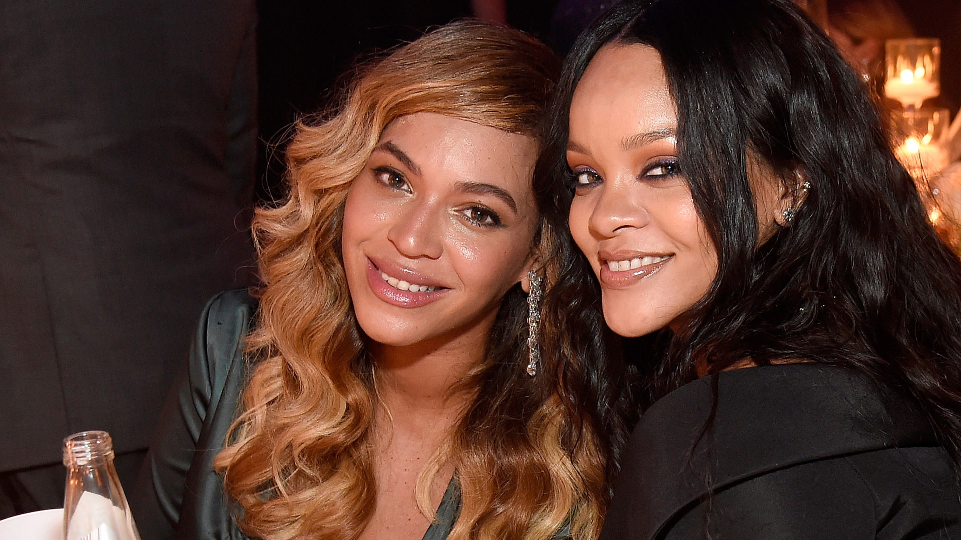 Beyoncé and Rihanna smiling at an event, sitting close together. Beyoncé has wavy hair, and Rihanna has long, straight hair.