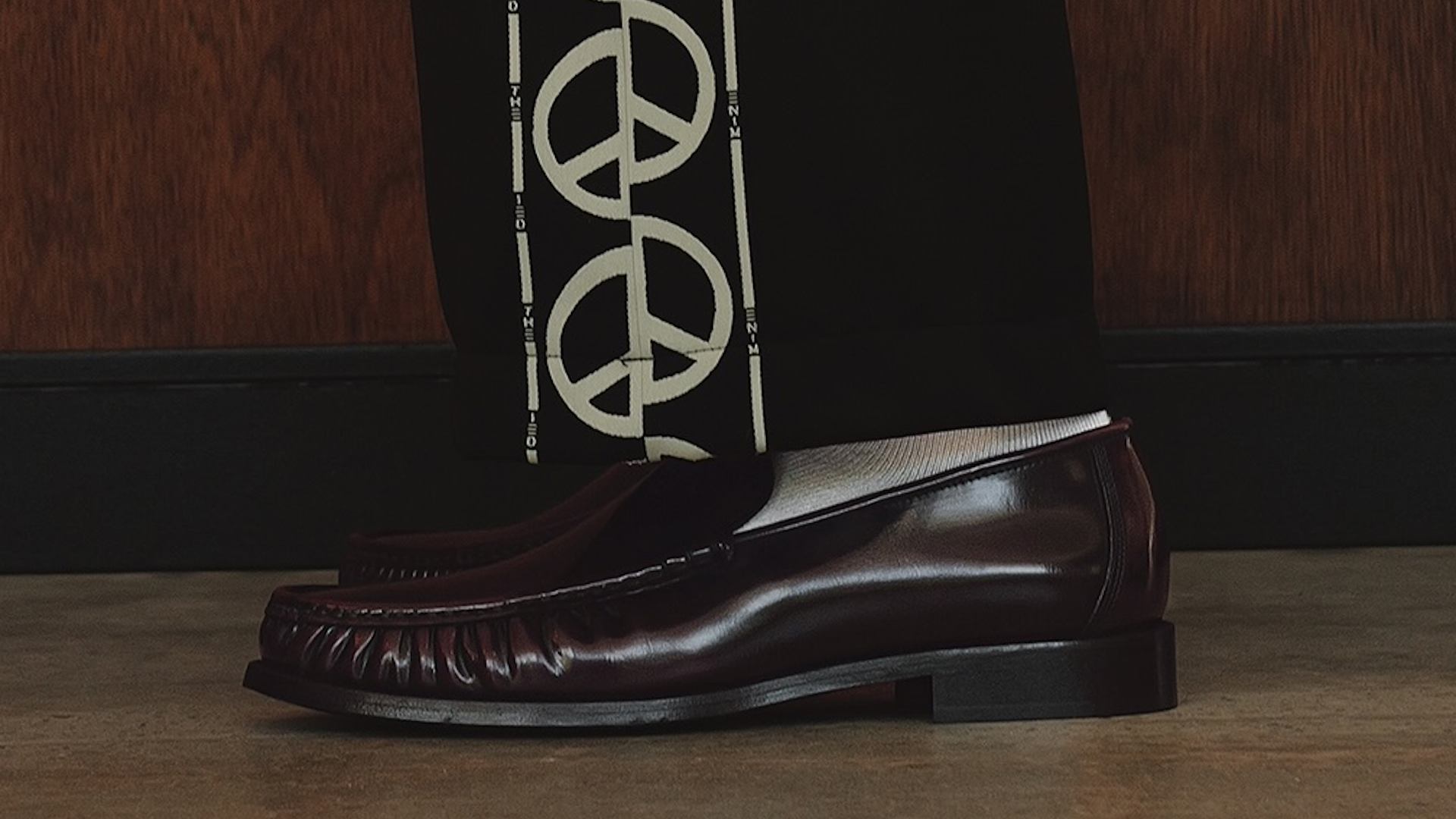 Brown leather loafers with white socks, worn with black pants featuring a bold white peace sign pattern, on a wooden floor.