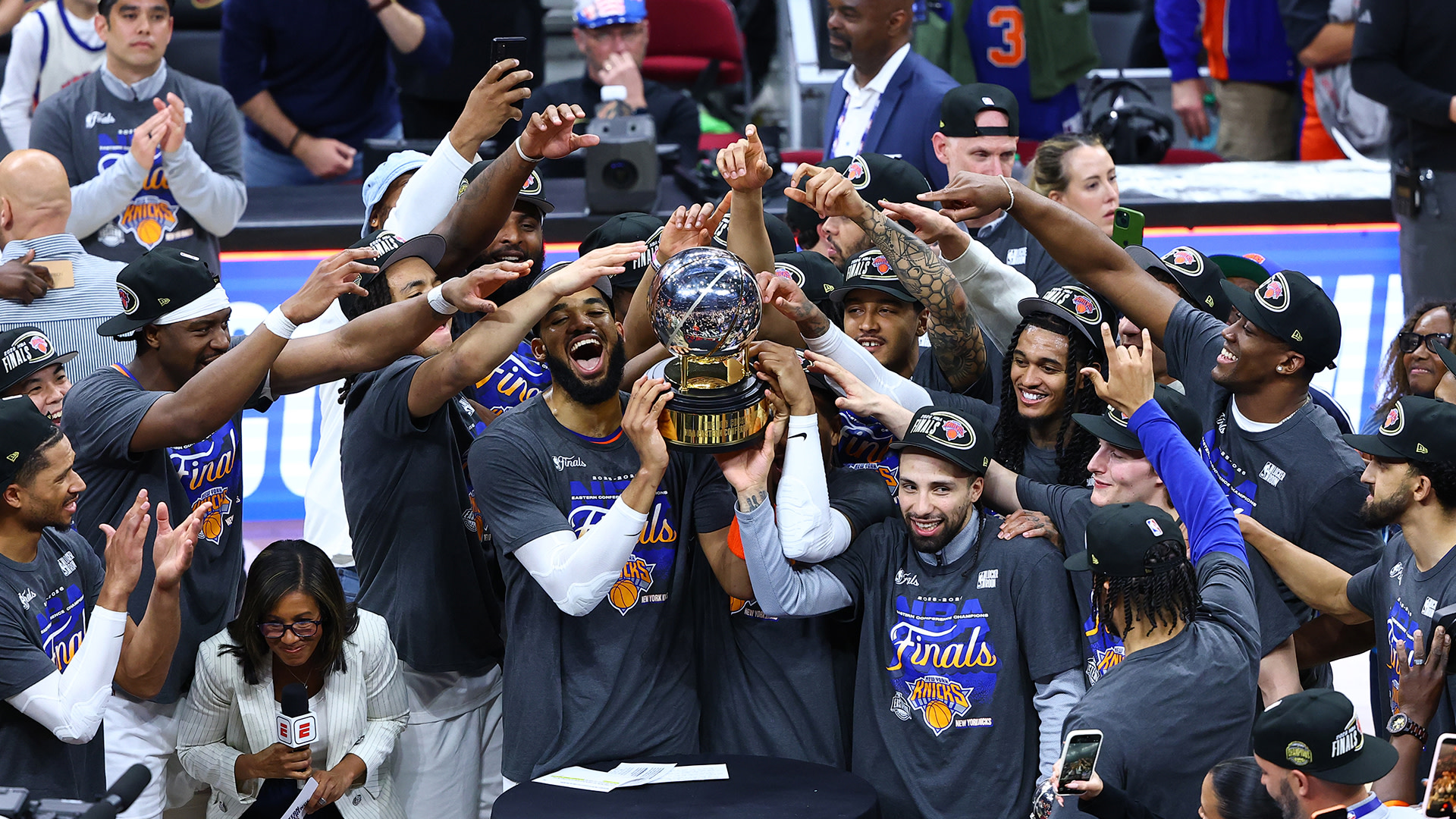 The New York Knicks celebrate, holding a trophy. Players wear matching "Finals" shirts and caps, with media and fans capturing the moment.