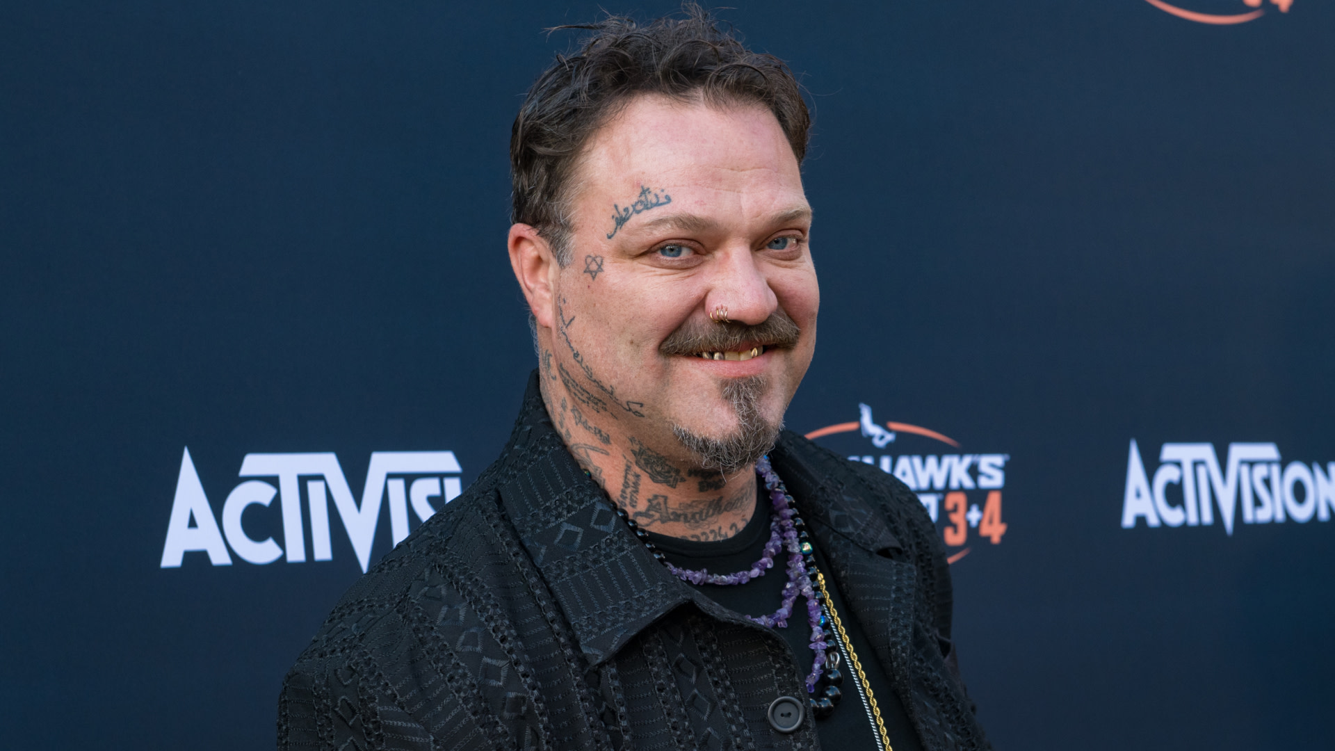 Bam Margera with facial tattoos and a beard smiles at an event, standing in front of a backdrop with logos.