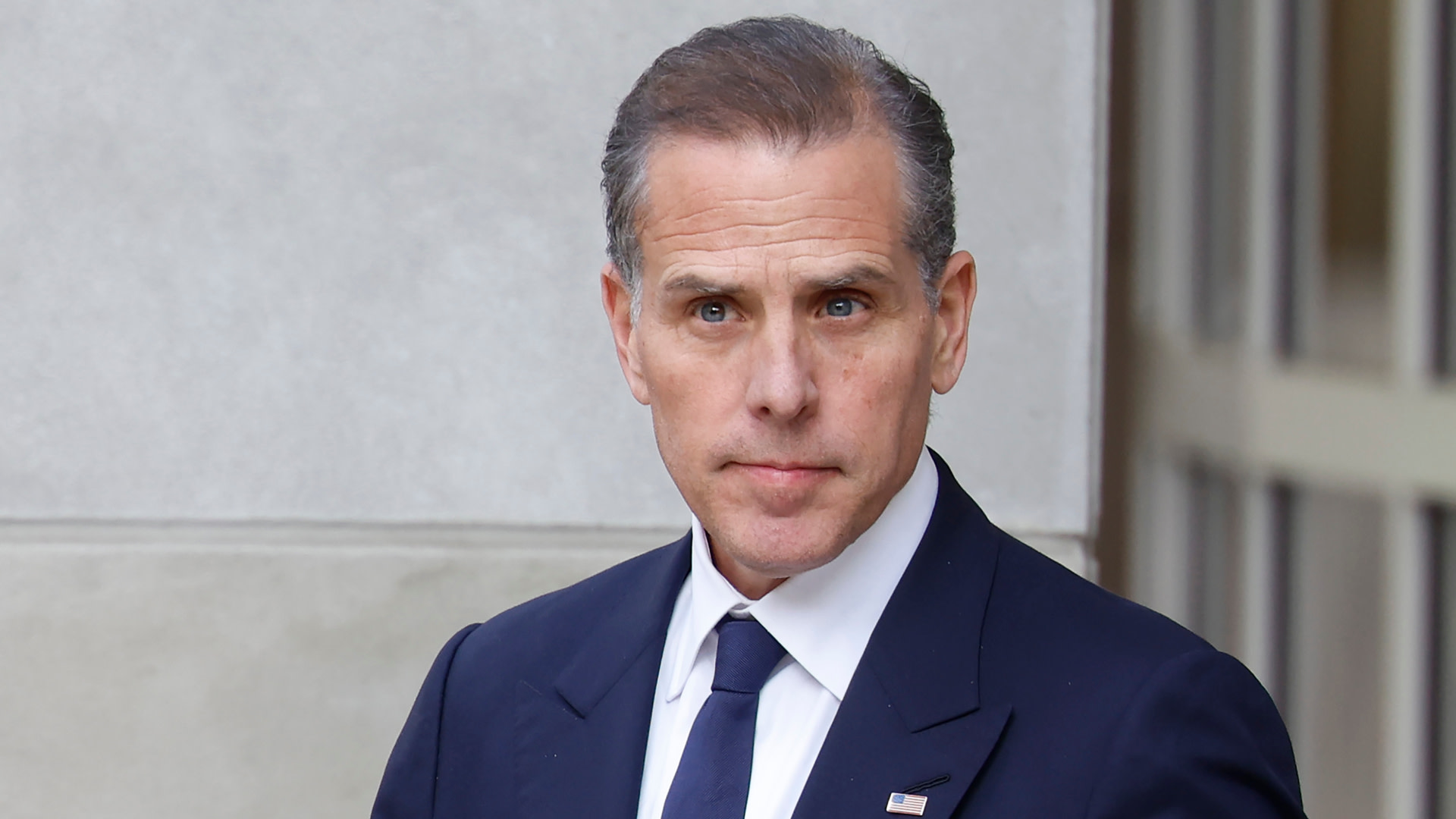 Hunter Biden in a suit and tie stands outside a building, looking directly at the camera.