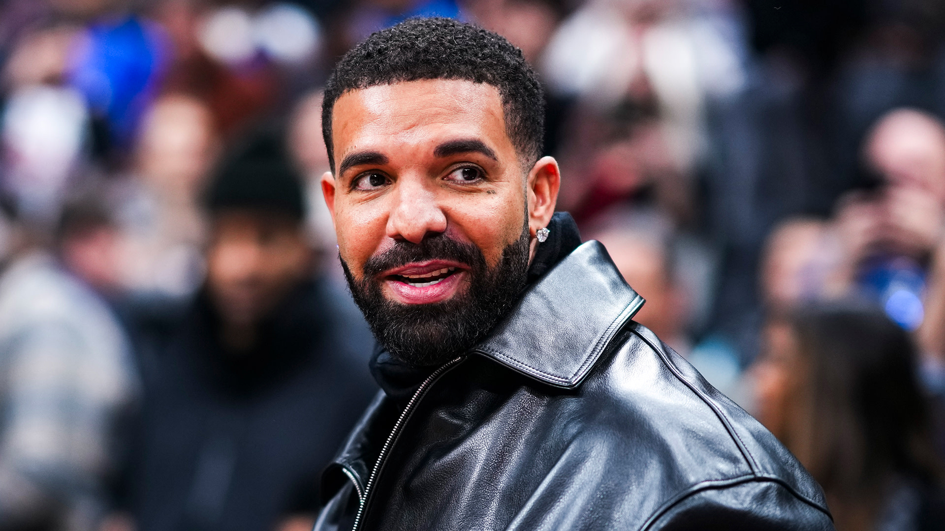 Drake in a black leather jacket, smiling, with a blurred crowd in the background.