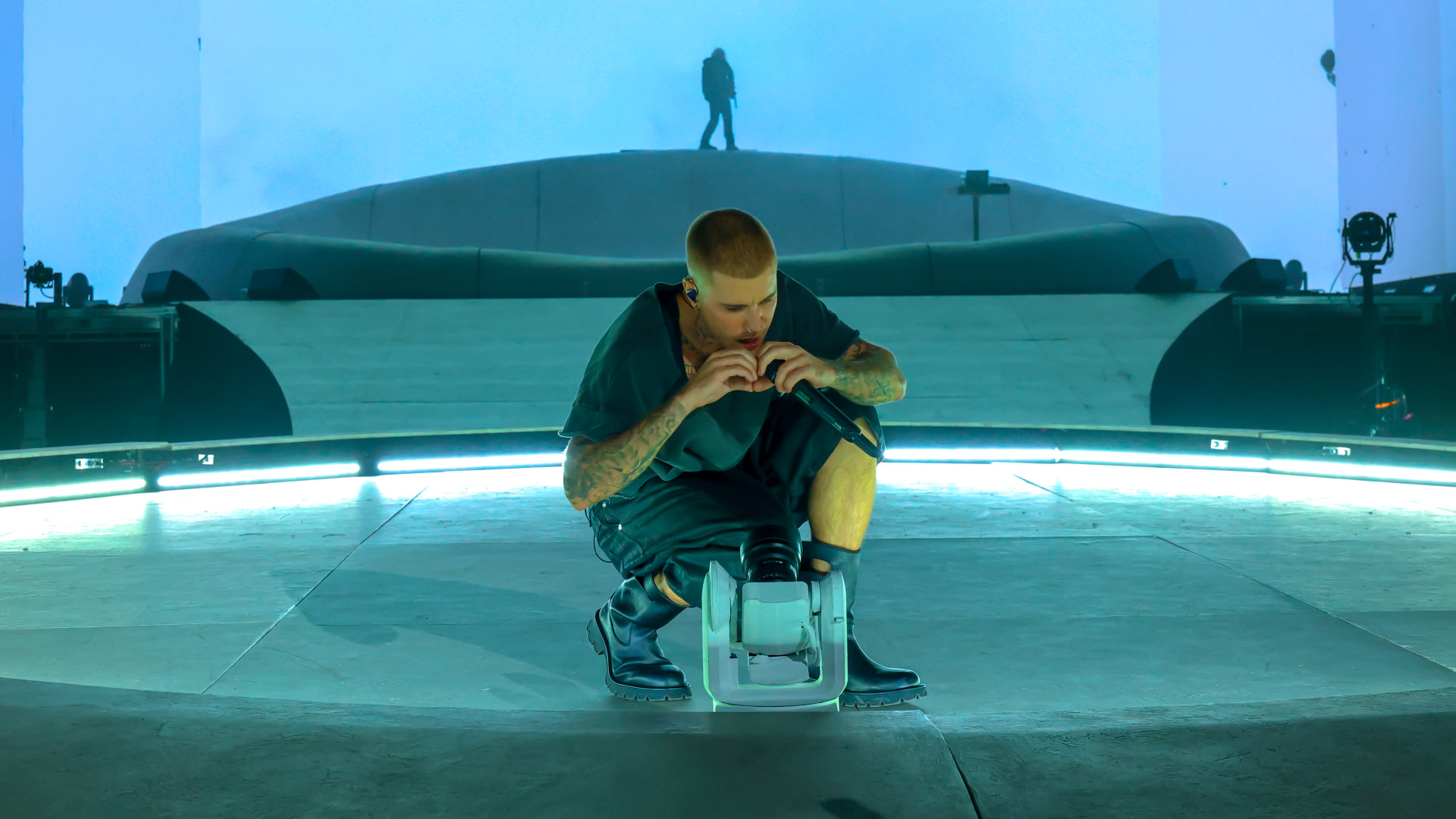 Justin Bieber crouches on stage with a microphone, a silhouette in the background. The stage is illuminated with blue lighting.