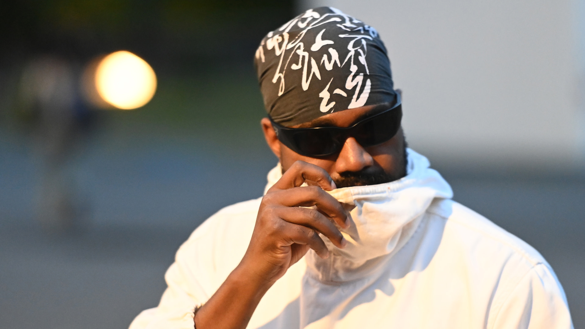 Ye wearing a headscarf with white patterns, sunglasses, and a white jacket, partially covering their face with their hand.