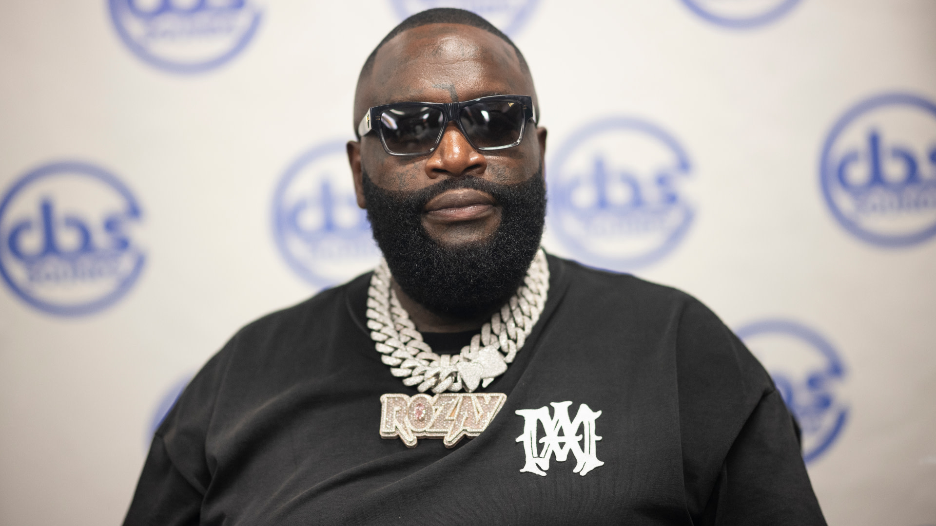 Rick Ross wearing sunglasses and a large chain, standing in front of a backdrop with logos.