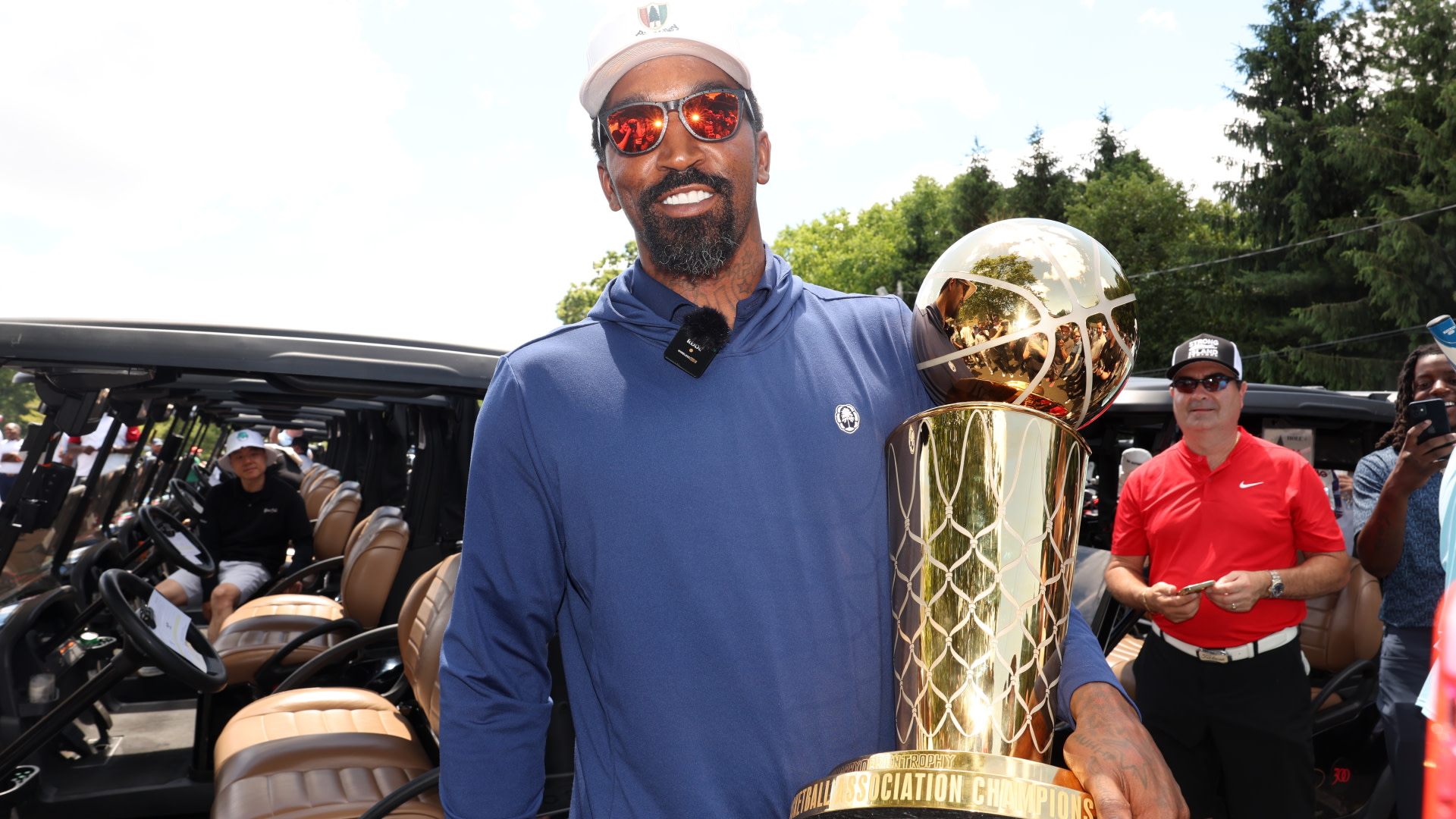 JR Smith in sunglasses and a blue shirt holds a large trophy outdoors, with people and golf carts in the background.