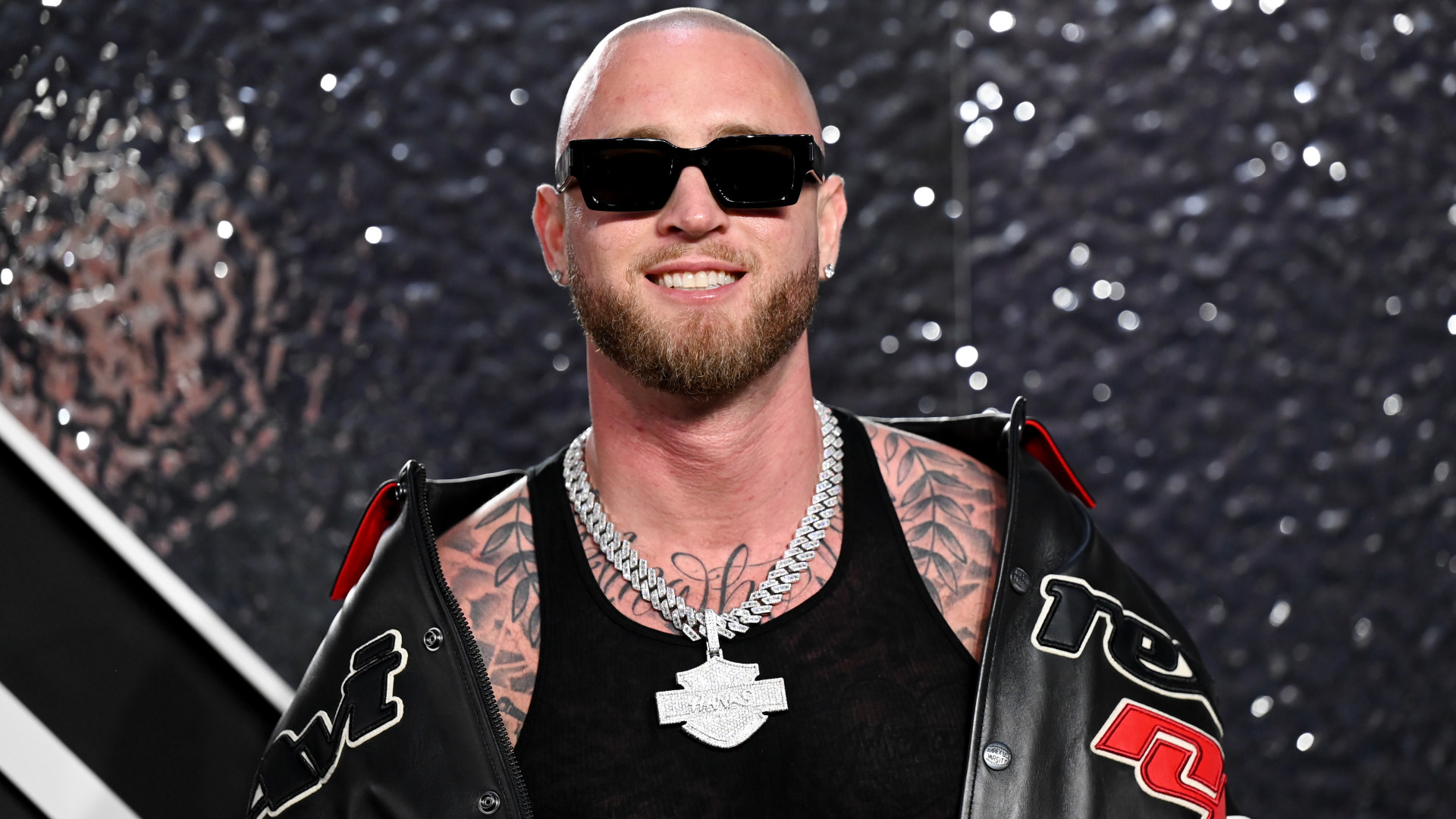 Chet Hanks with a beard and sunglasses, wearing a black jacket and chain necklace, smiles against a textured background.