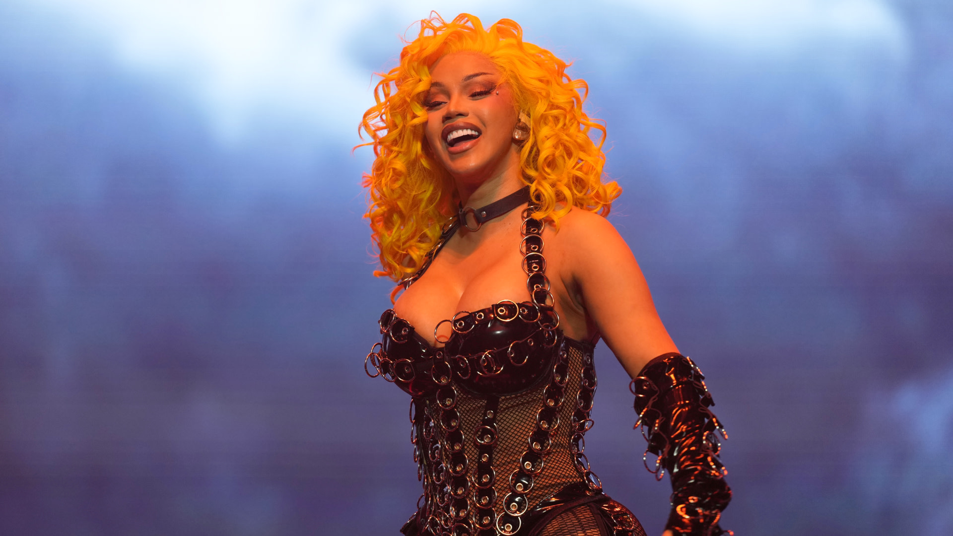 Cardi B with bright yellow curly hair, wearing a black corset with silver rings, smiles on stage against a blue background.