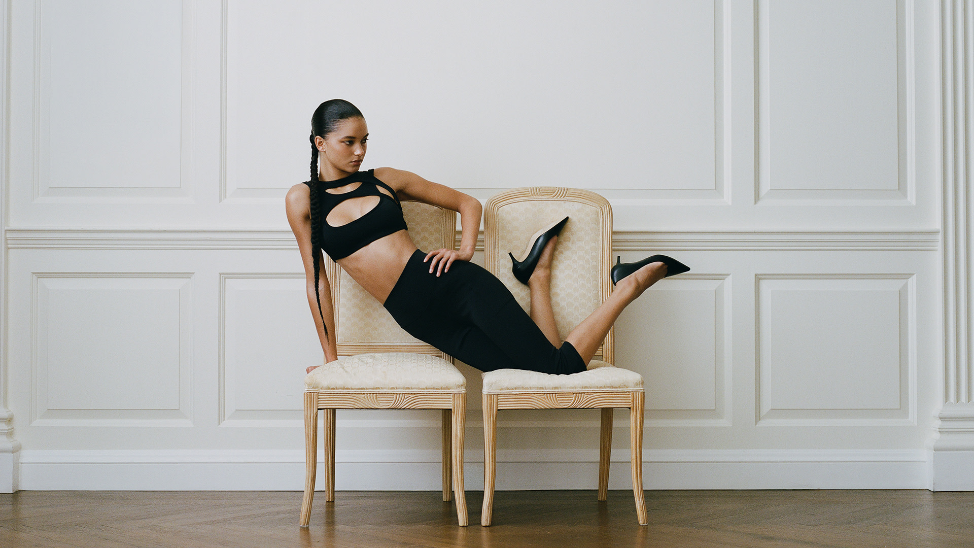 A woman in a black outfit and heels poses elegantly on two chairs against a white paneled wall.