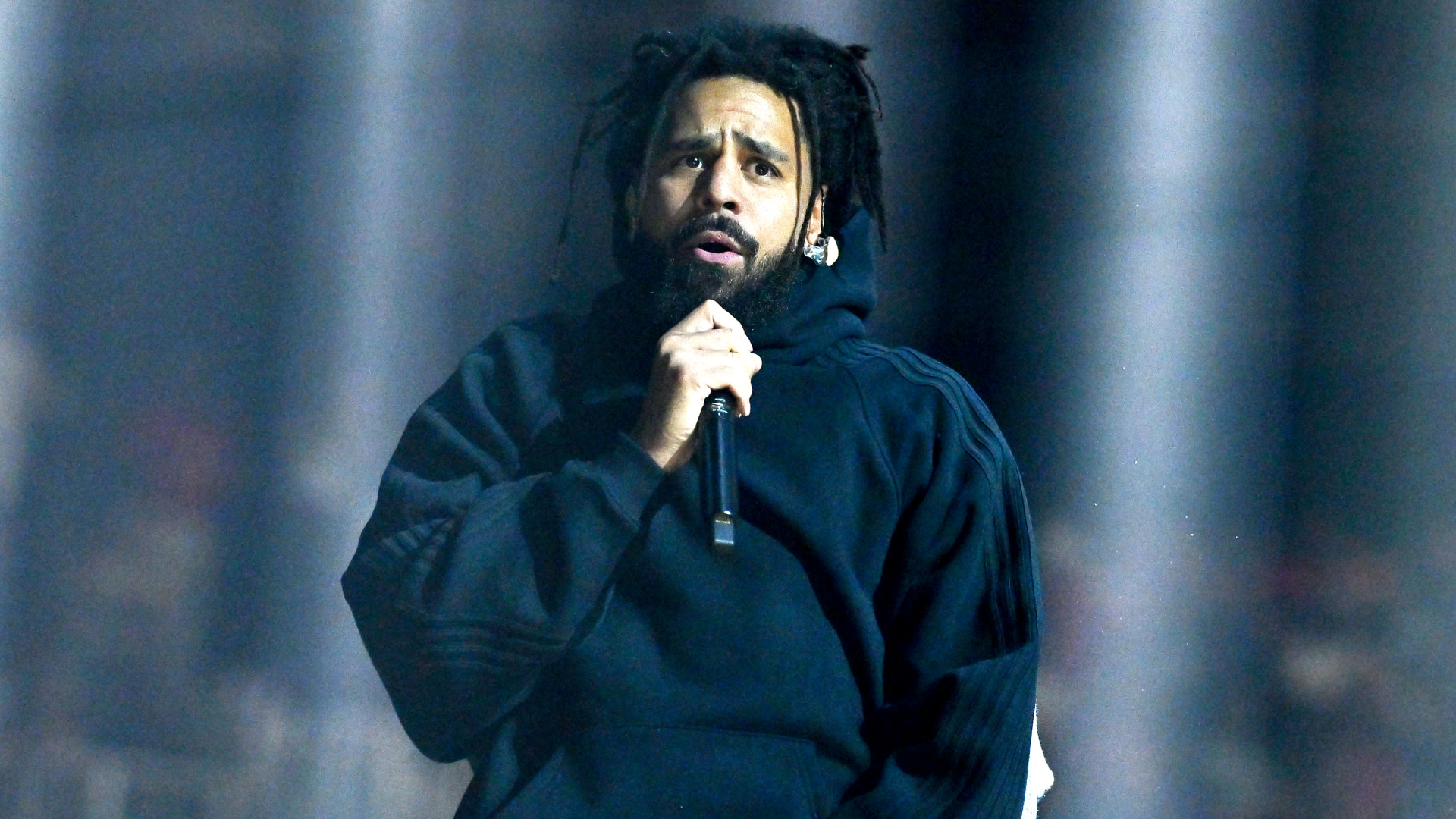 J. Cole performing on stage, wearing a dark hoodie and holding a microphone, with an expressive look.