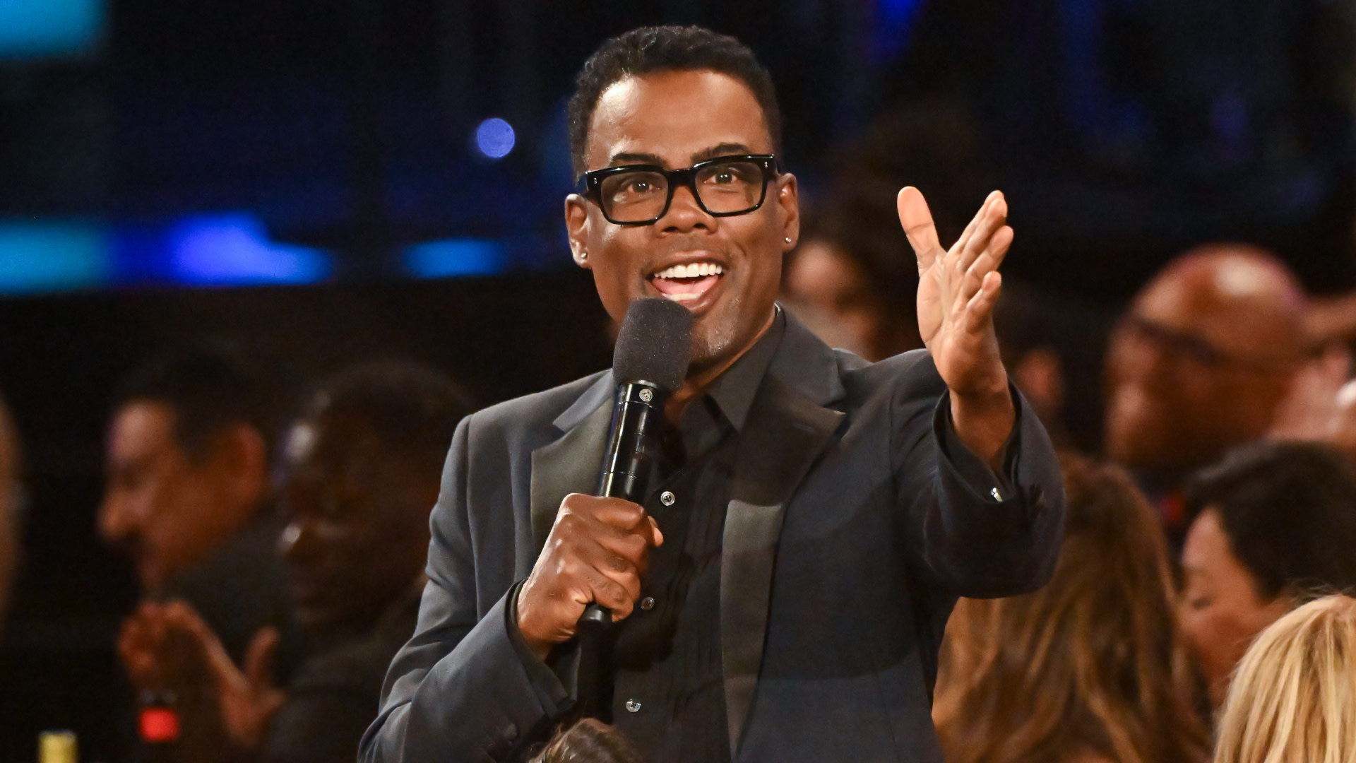 Comedian Chris Rock speaking into a microphone, wearing glasses and a suit, gesturing with his hand at an event.