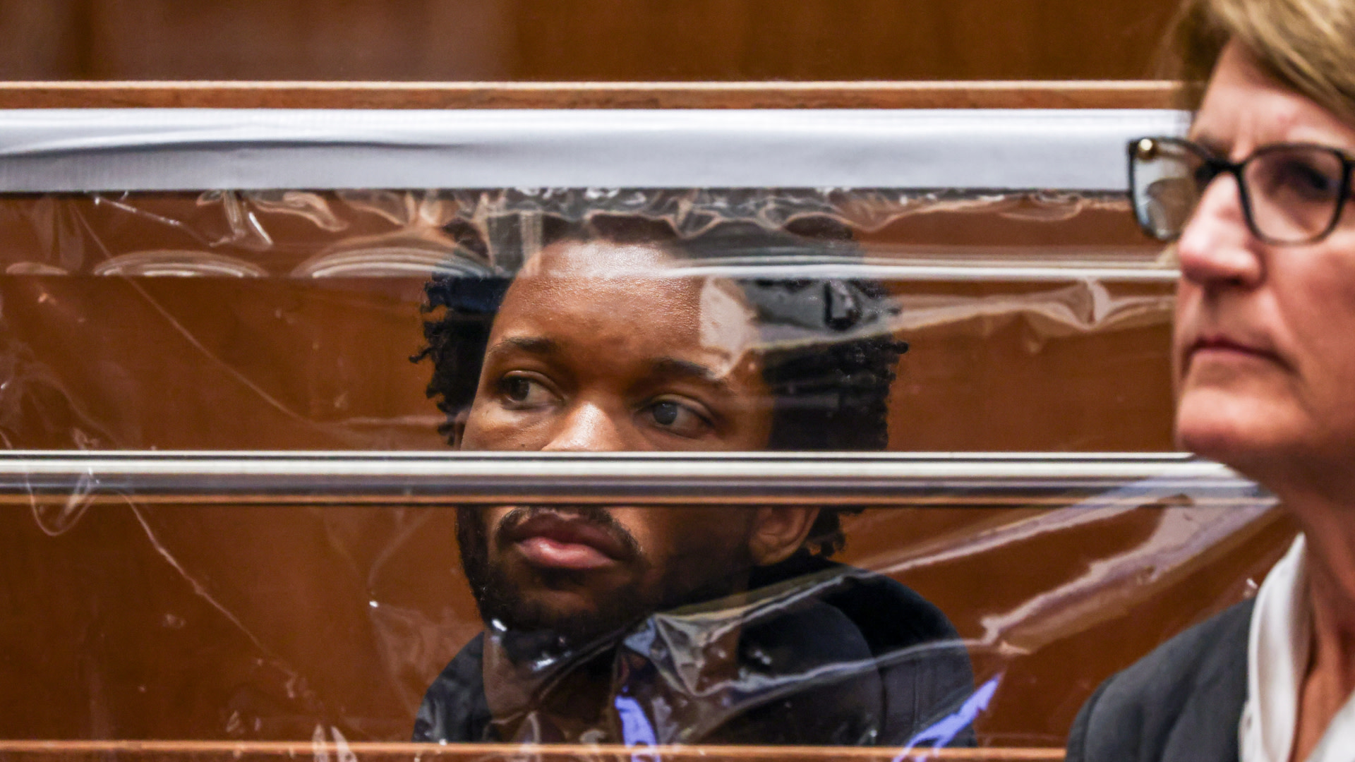 D4vd behind a transparent barrier looks forward, while a woman with glasses sits in the foreground.