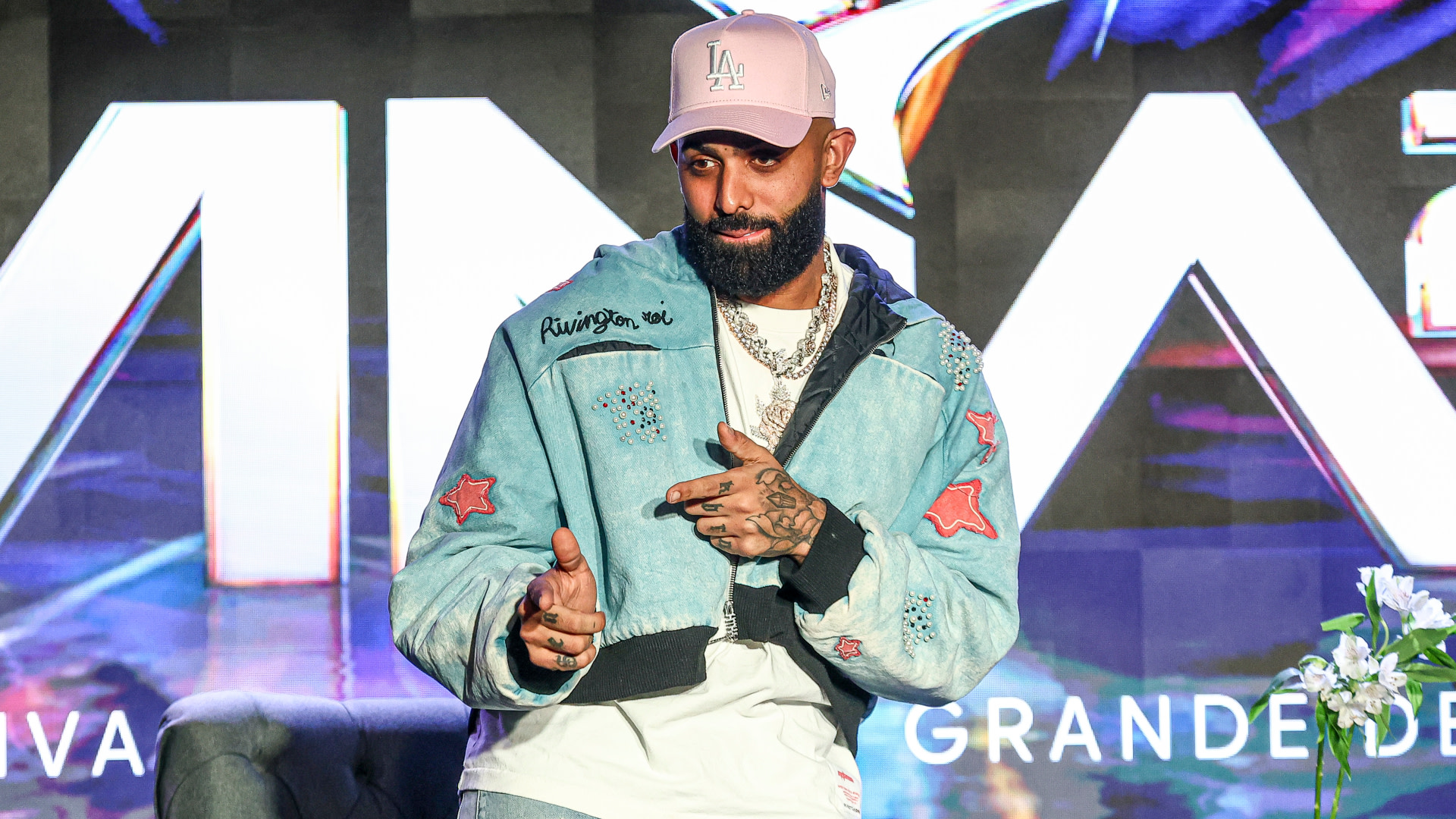 Eladio Carrión with a beard wearing a light blue jacket and a pink LA cap stands in front of a colorful background with large text.