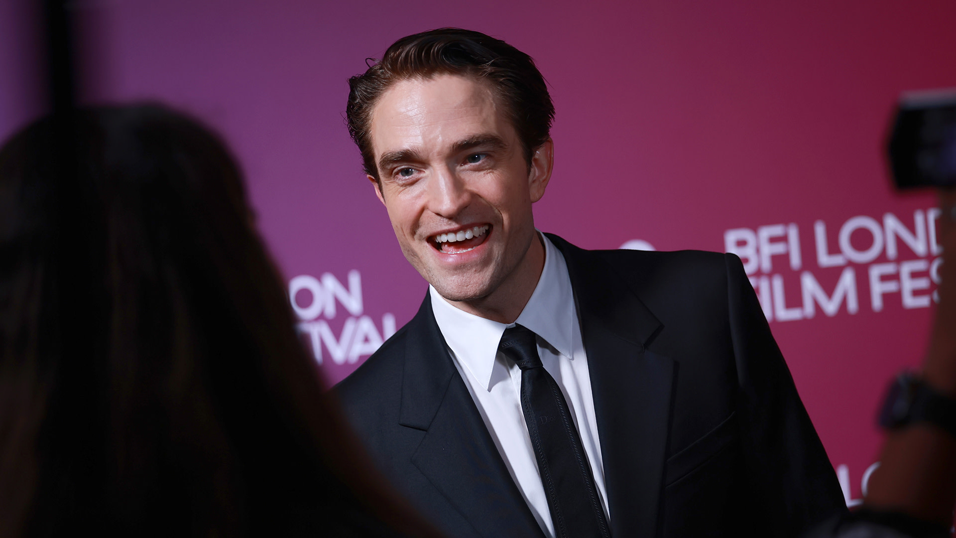Robert Pattinson at the BFI London Film Festival, wearing a black suit and tie, smiling against a red backdrop.