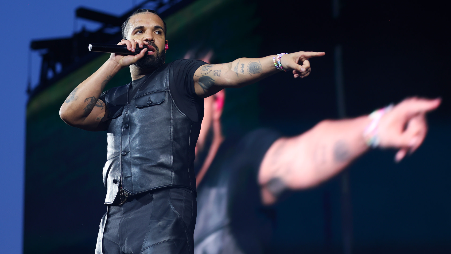 Drake performing on stage, wearing a black leather vest, holding a microphone, and pointing into the crowd.