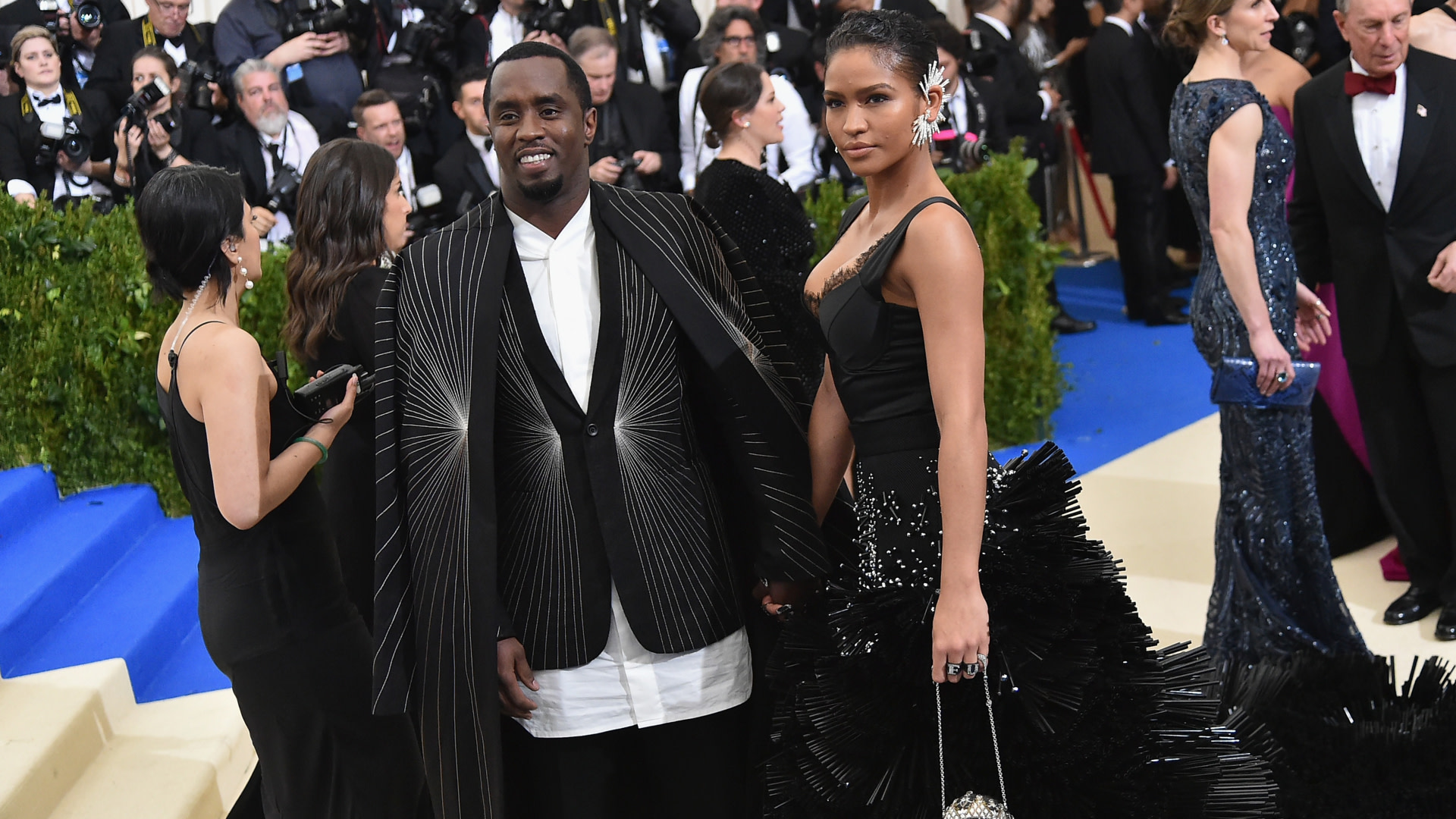 Sean "Diddy" Combs in a stylish black and white outfit, and Cassie in a black gown, pose at a formal event with a crowd in the background.
