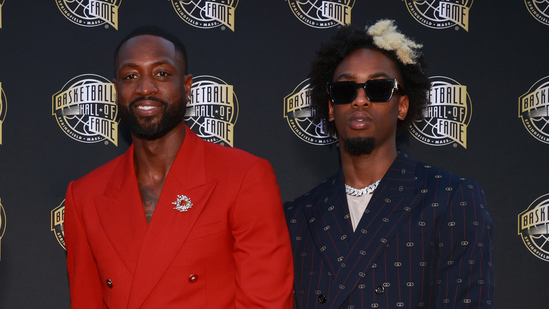 Dwyane Wade in a red suit and son Zaire Wade in a striped suit with sunglasses at the Basketball Hall of Fame event.