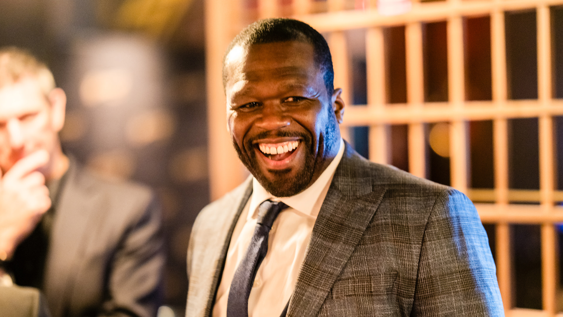 50 Cent, wearing a gray suit and tie, smiles broadly in a warmly lit setting.