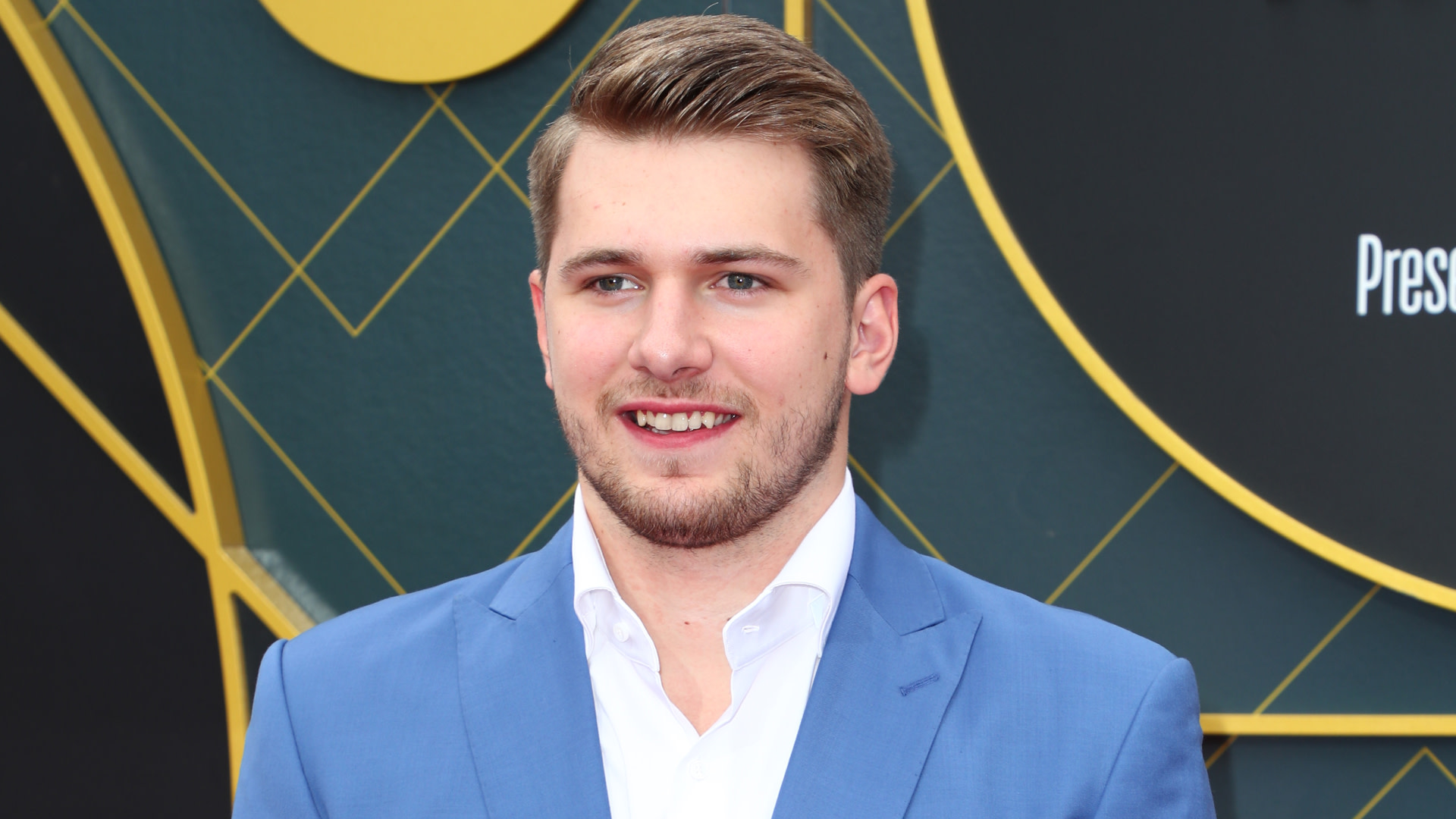 Luka in a blue suit and white shirt smiles at an event, standing against a decorative background.