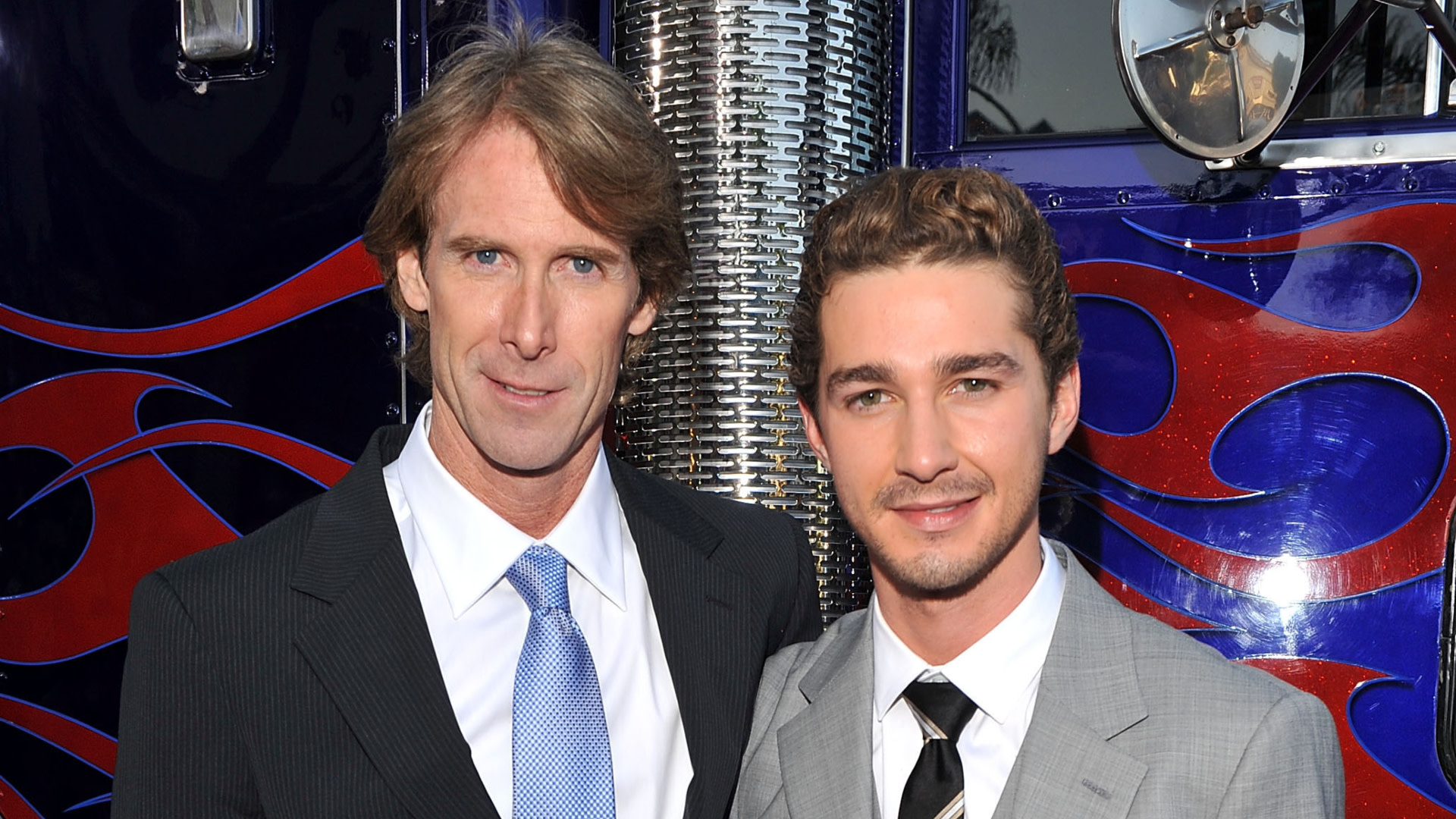 Michael Bay and Shia LaBeouf pose in front of a truck. Bay wears a dark suit, and LaBeouf a light gray suit with a striped tie.