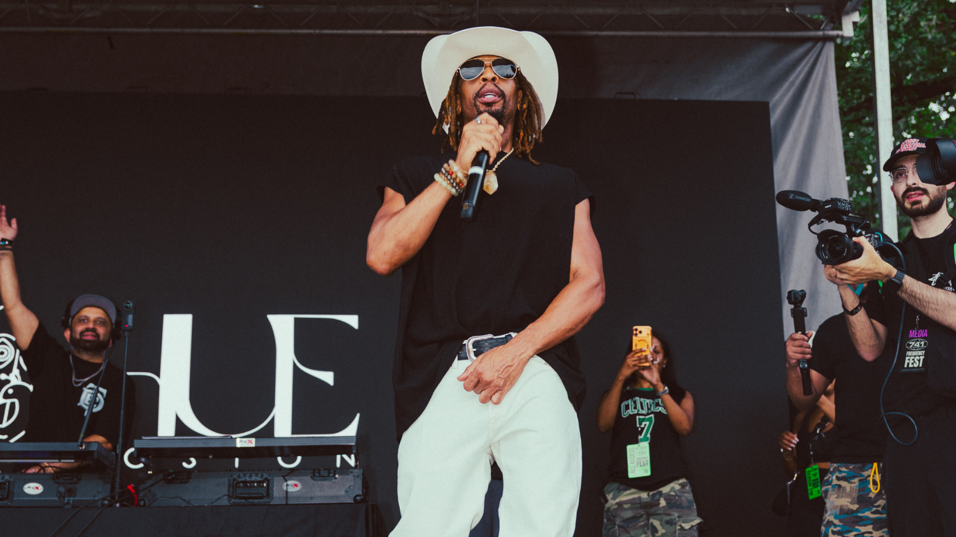 Lil Jon wearing a white hat and sunglasses sings on stage, with a DJ and cameramen in the background.